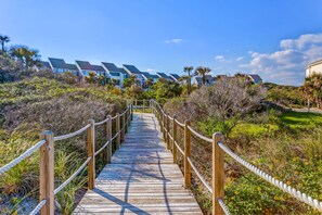 A wooden boardwalk leading to a row of modern buildings with sloped roofs, surrounded by dense vegetation and a clear sky. - Renovated Condo With Easy Beach Access, Balcony, Pool & Spectacular Ocean Views (Fernandina Beach)