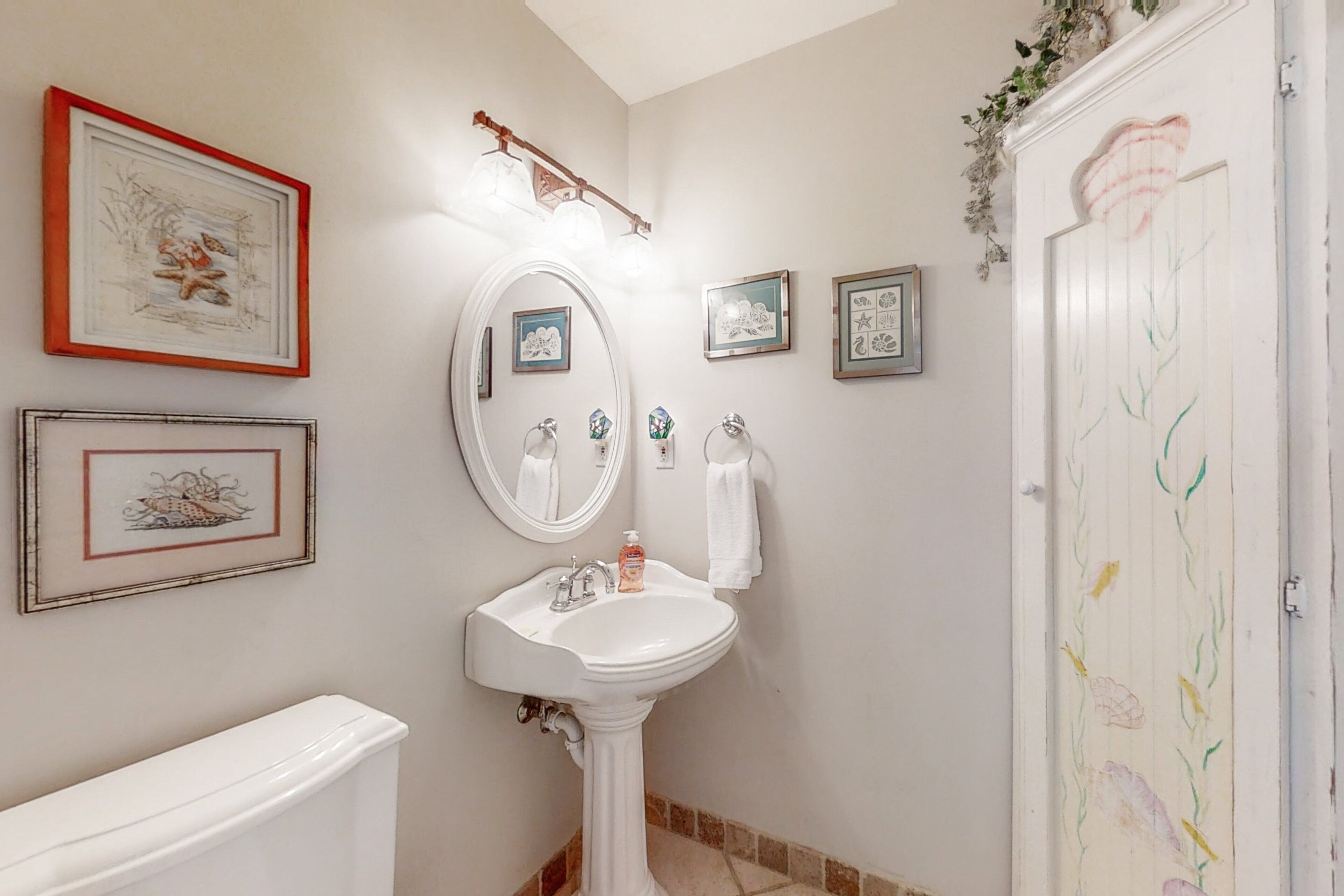 A bathroom with a pedestal sink, a large mirror, framed artwork on the walls, and a toilet.