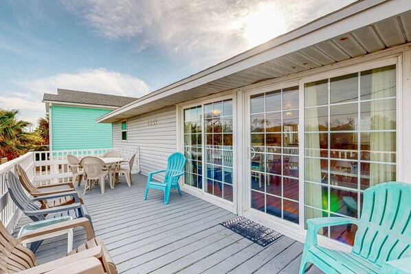 A covered outdoor deck with chairs and a table, large windows, and a teal-colored building. - Serene 2-level Home With Wifi & Deck - Walk to Beach, 1 dog Welcome (St. Augustine)