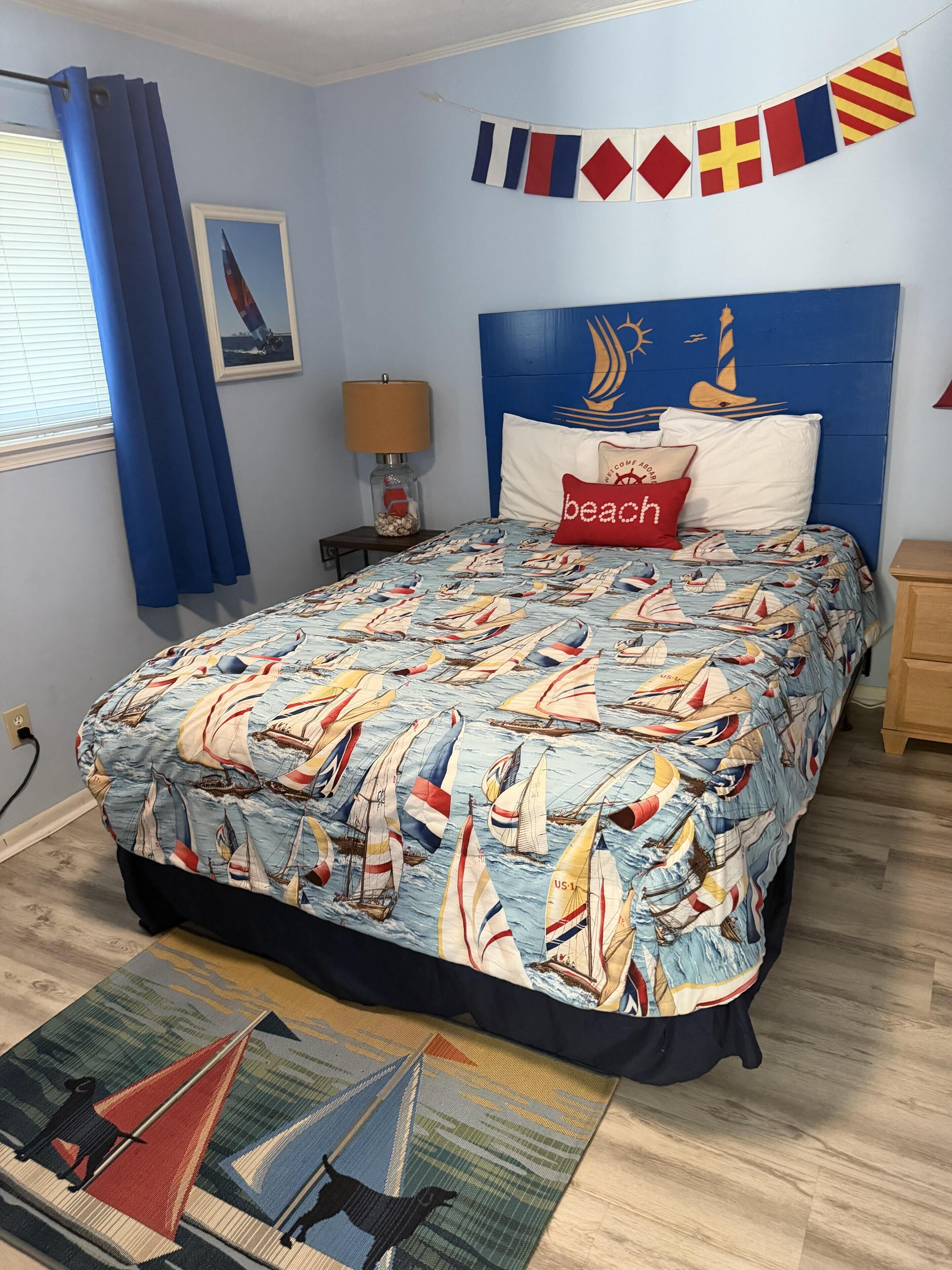 A bedroom with a bed featuring a nautical-themed bedspread, a headboard with a sailboat mural, a wall-mounted flag display, and a small table with a lamp.
