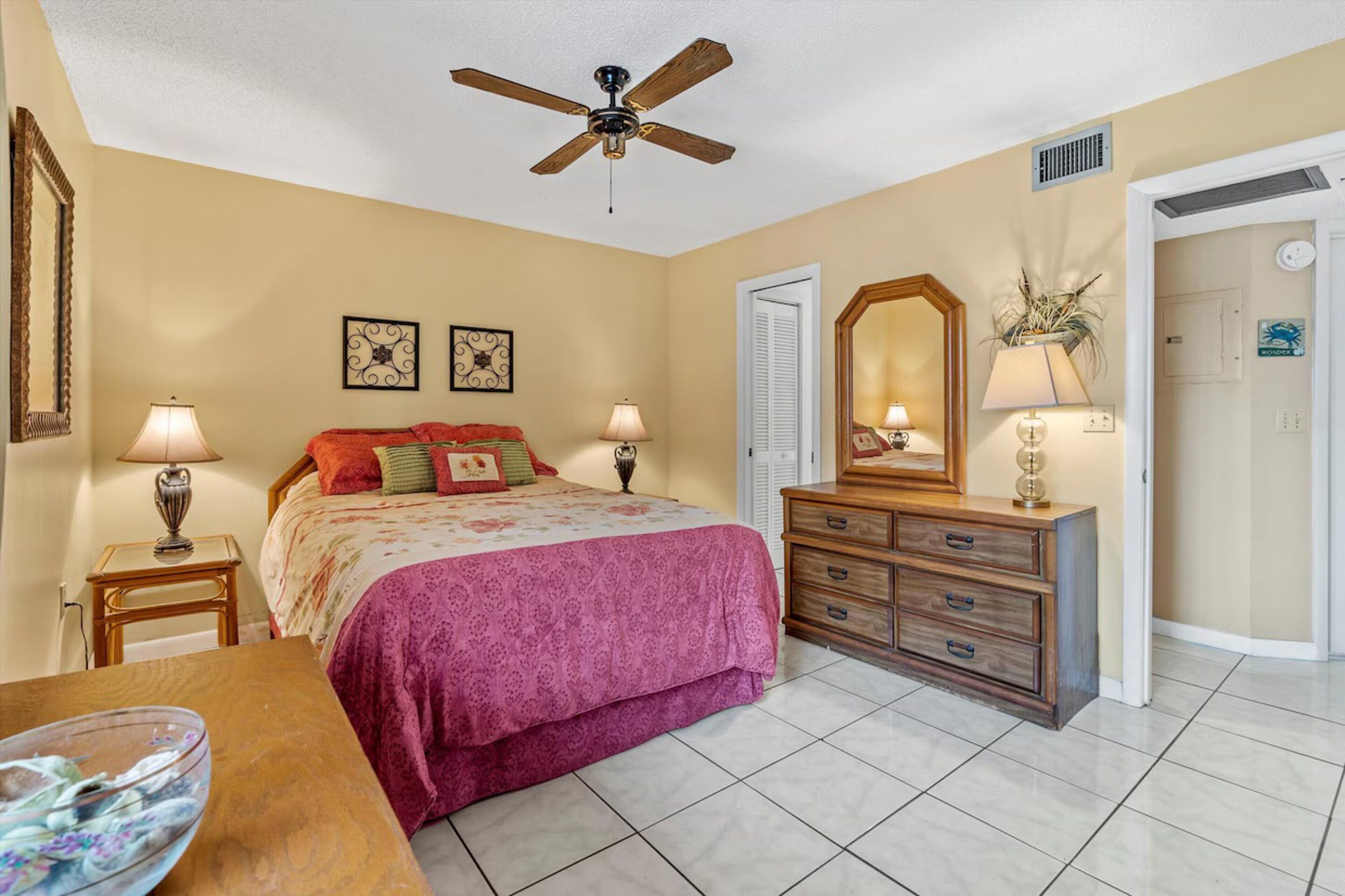 Une chambre à coucher comprenant un lit, une commode, deux tables de chevet, un ventilateur de plafond et une porte donnant sur une autre pièce.