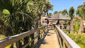 A wooden boardwalk leading to a building with a tiled roof, surrounded by palm trees and greenery. - Spacious Condo With Private Balcony and Close Beach Access (St. Augustine)