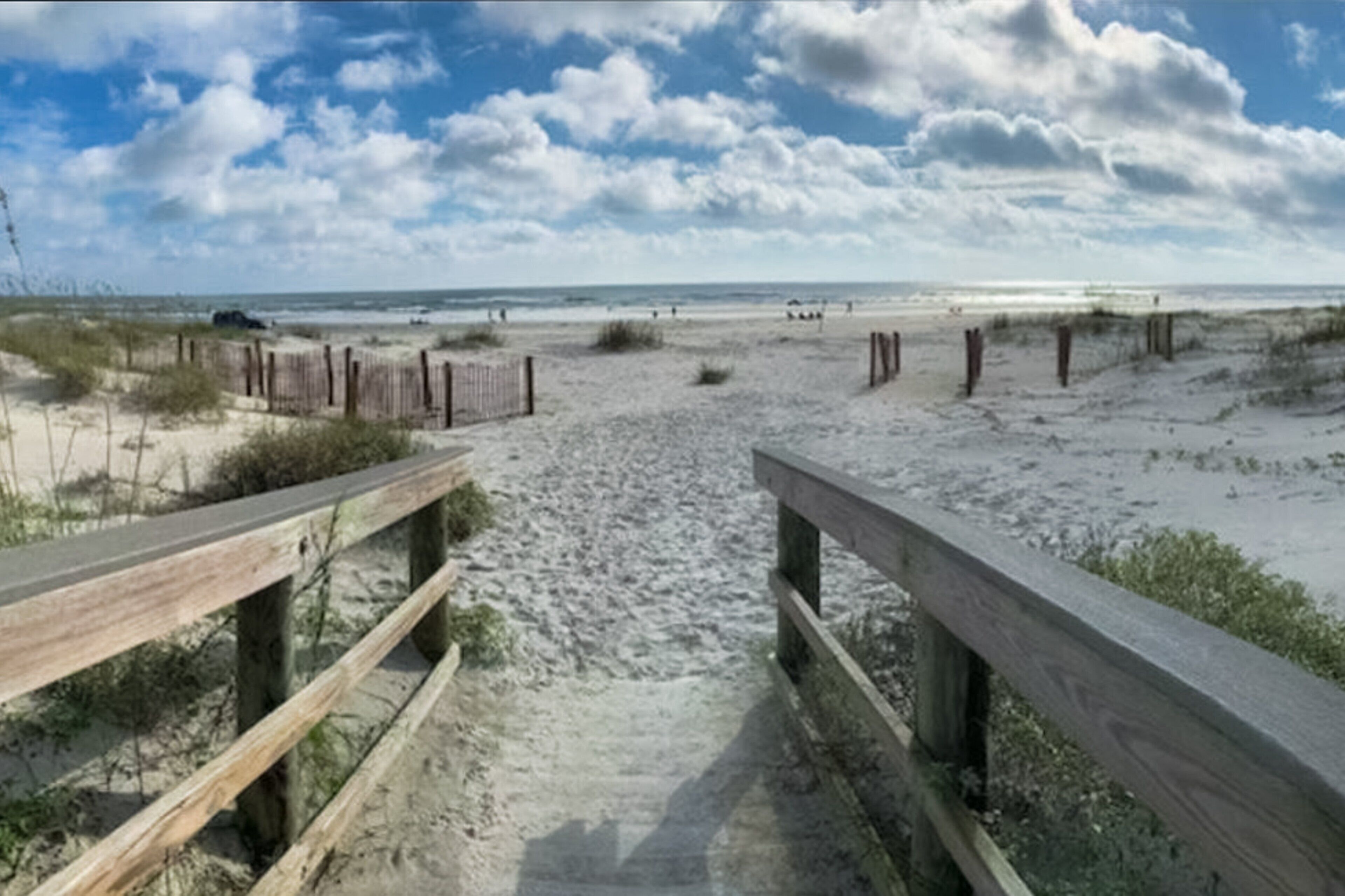 Une plage de sable avec des rambardes en bois menant à l’eau, sous un ciel partiellement nuageux.
