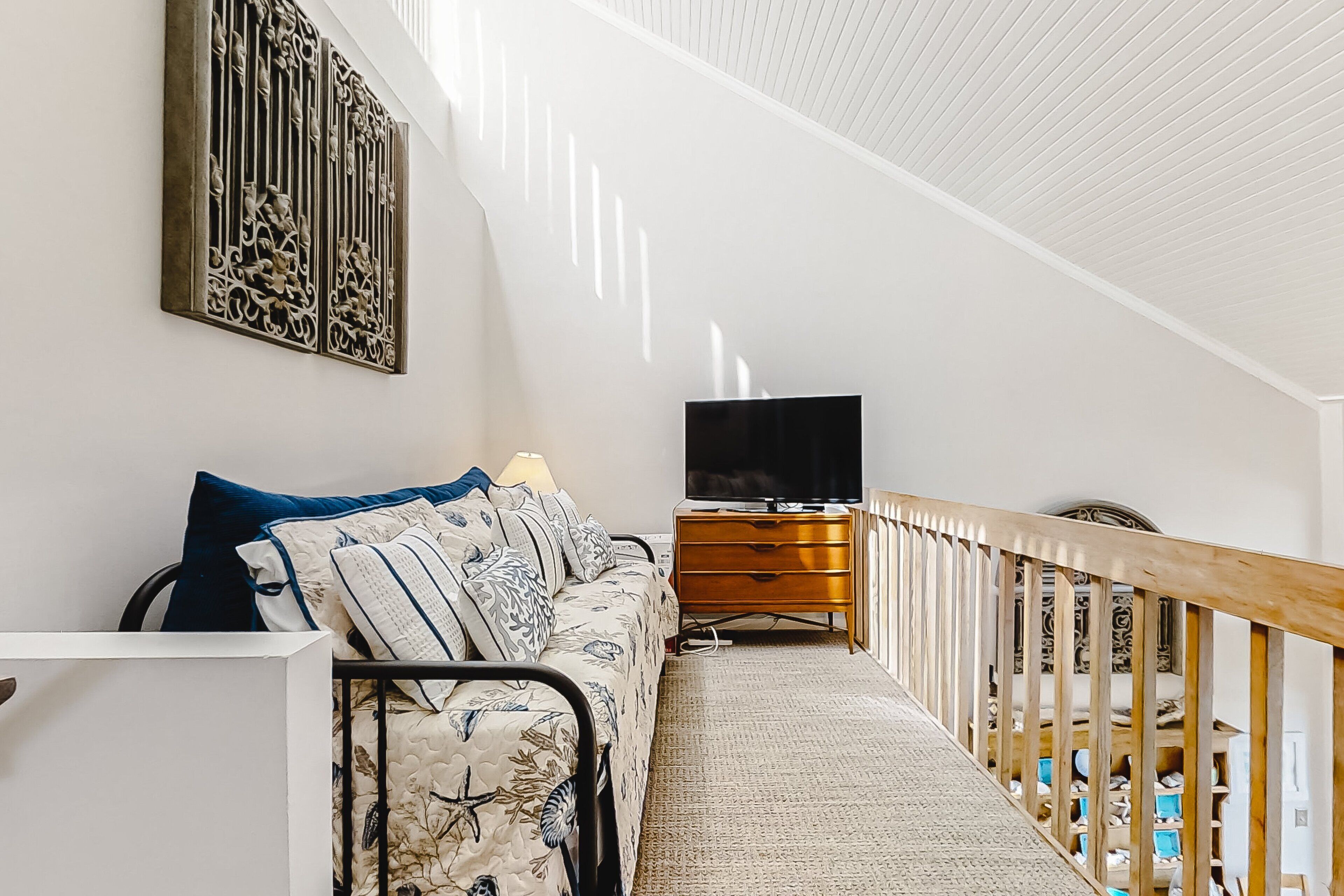 Un couloir étroit avec une rambarde en bois, un canapé orné de coussins à motifs, un téléviseur posé sur un meuble en bois et un élément décoratif mural.