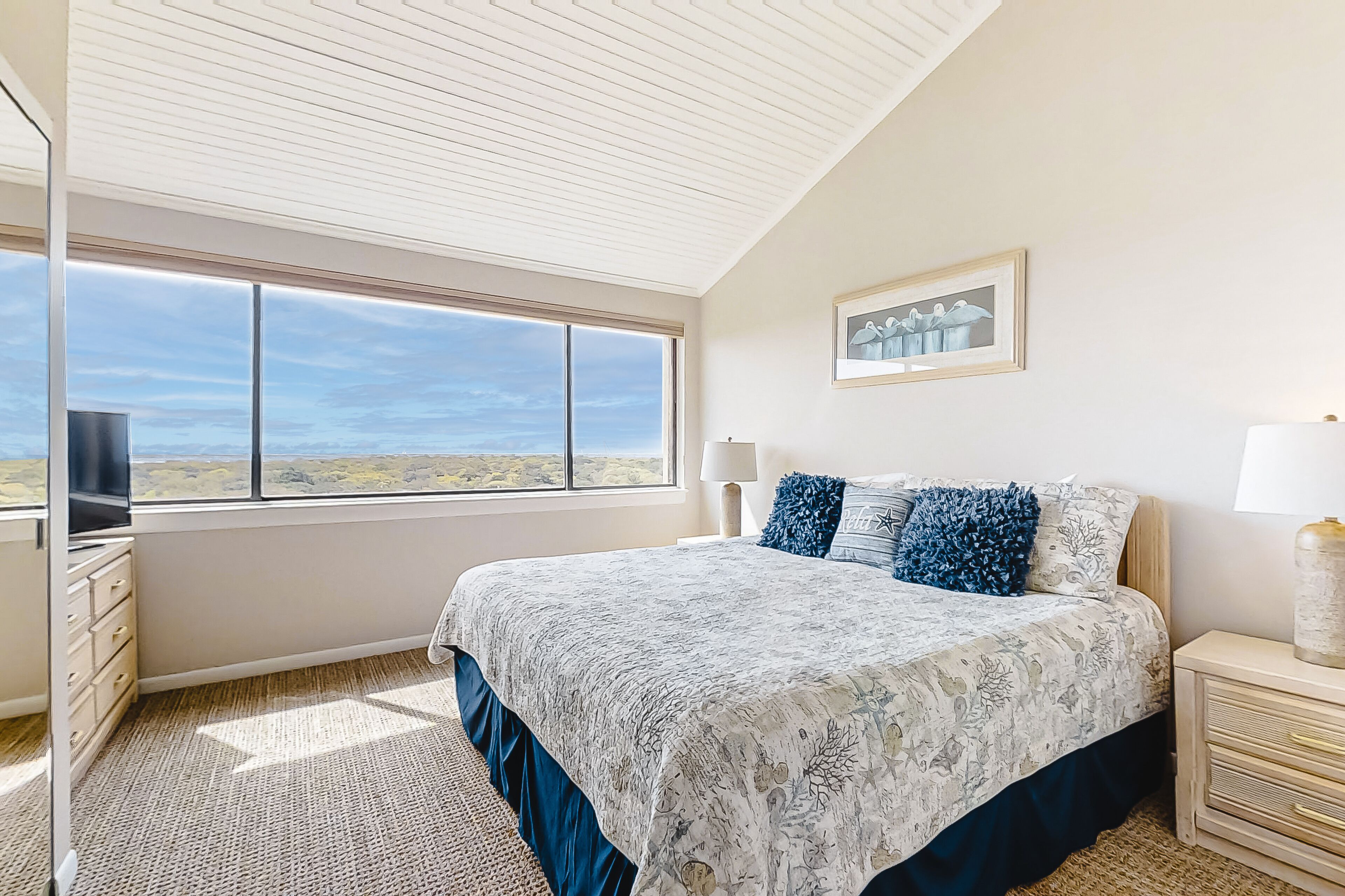 A bedroom with a large window, a bed with patterned bedding, a nightstand, and a wall-mounted lamp.