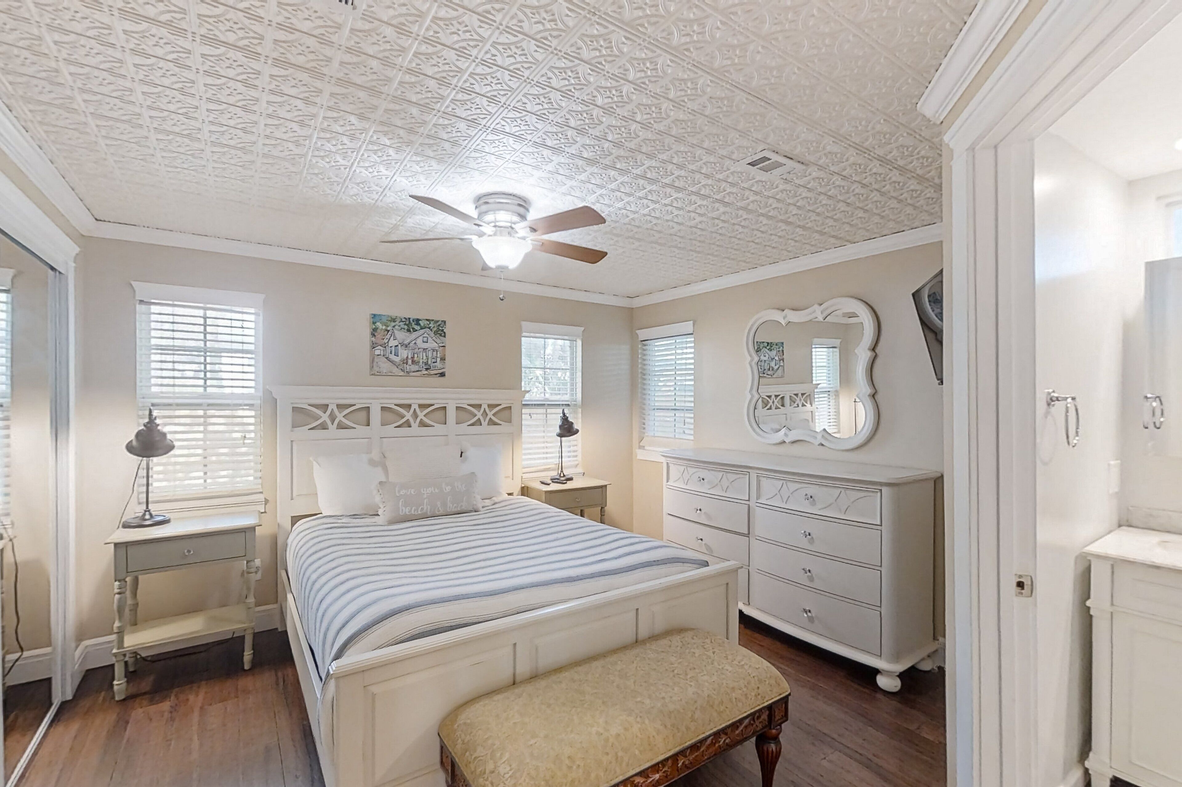 Quarto com cama, mesas de cabeceira, cômoda e ventoinha no teto.