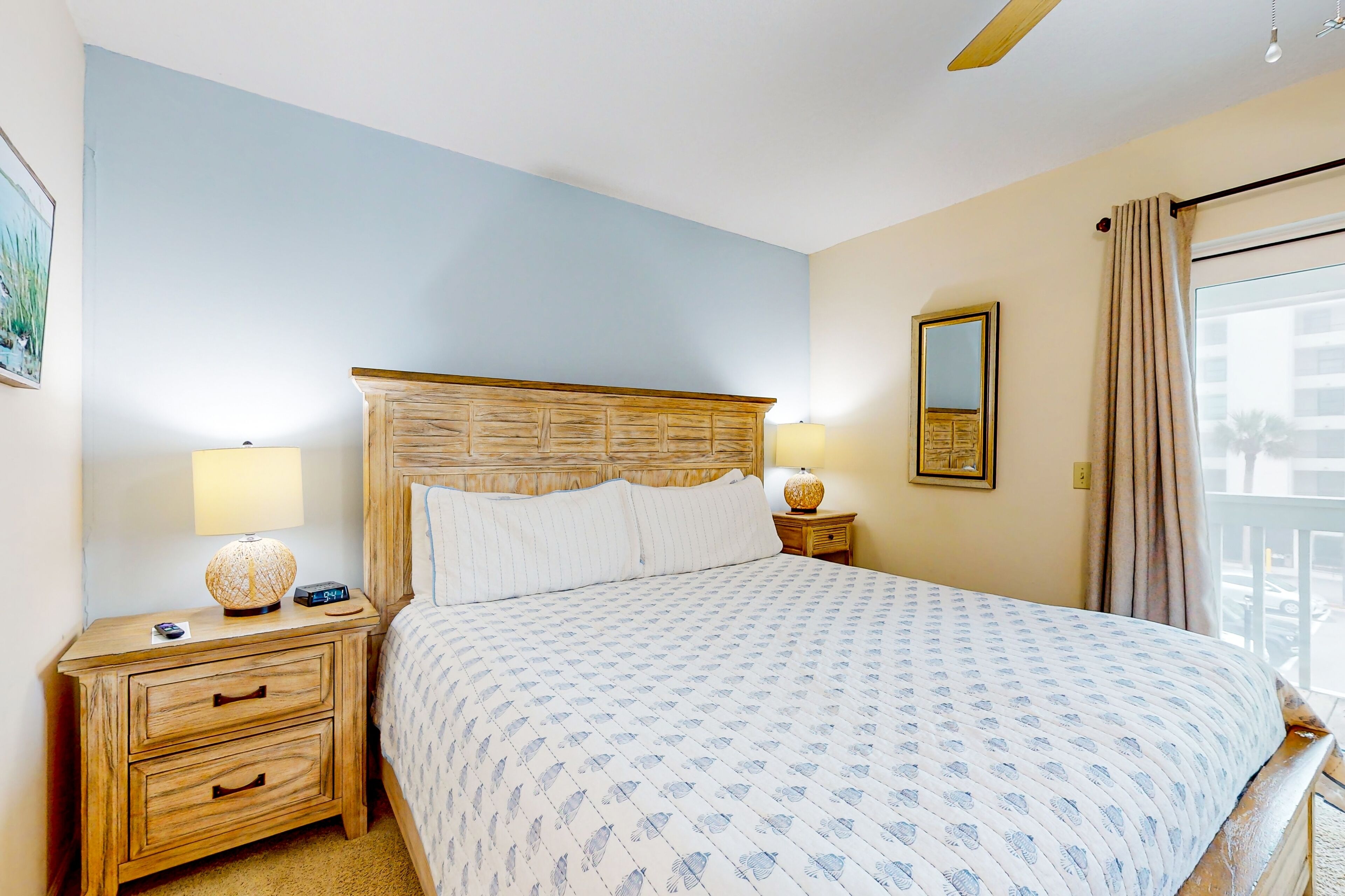 Un dormitorio con cama, cabecera de madera, mesitas de noche con lámparas, una mesita con cajones, un espejo y vistas al exterior.