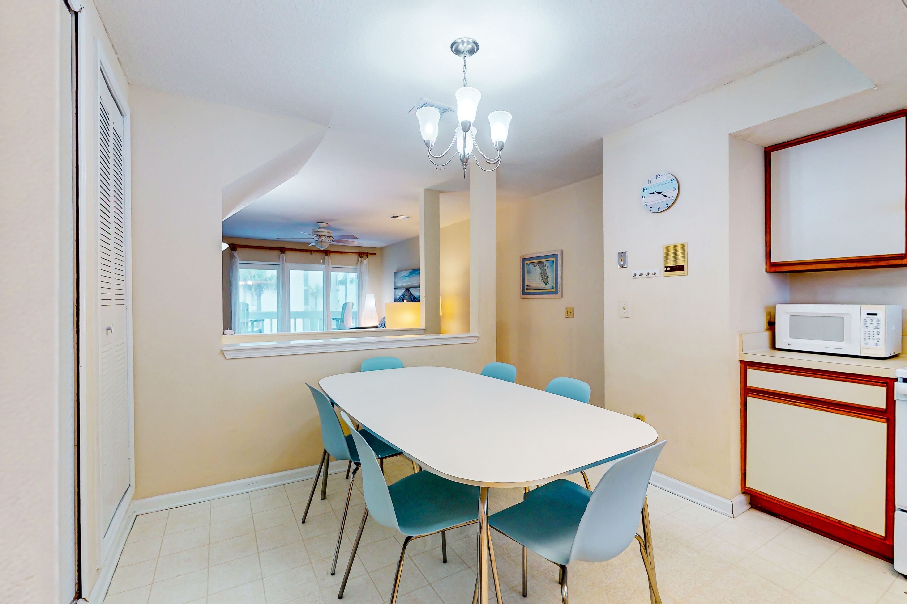 A dining area with a white table, blue chairs, a chandelier, a clock, and a microwave on a cabinet.
