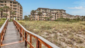 A wooden boardwalk leading to a beachfront residential complex with multiple balconies and a clear sky. - Ocean-view Penthouse With Pool, Balcony & Central AC - Snowbird-friendly (Fernandina Beach)