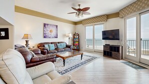 A spacious living room with a leather sofa, a white sofa, a coffee table, a ceiling fan, a TV, and a large window with a view. - Ocean-view Penthouse With Pool, Balcony & Central AC - Snowbird-friendly (Fernandina Beach)