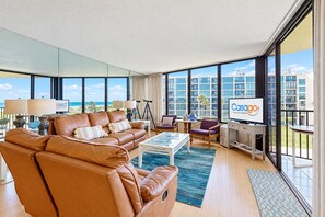 A modern living room with a leather sofa, a coffee table, and a TV. Large windows offer a view of the cityscape and a balcony. - Oceanfront Condo w/ Private Balcony & Stunning View - Boardwalk to Beach (St. Augustine)