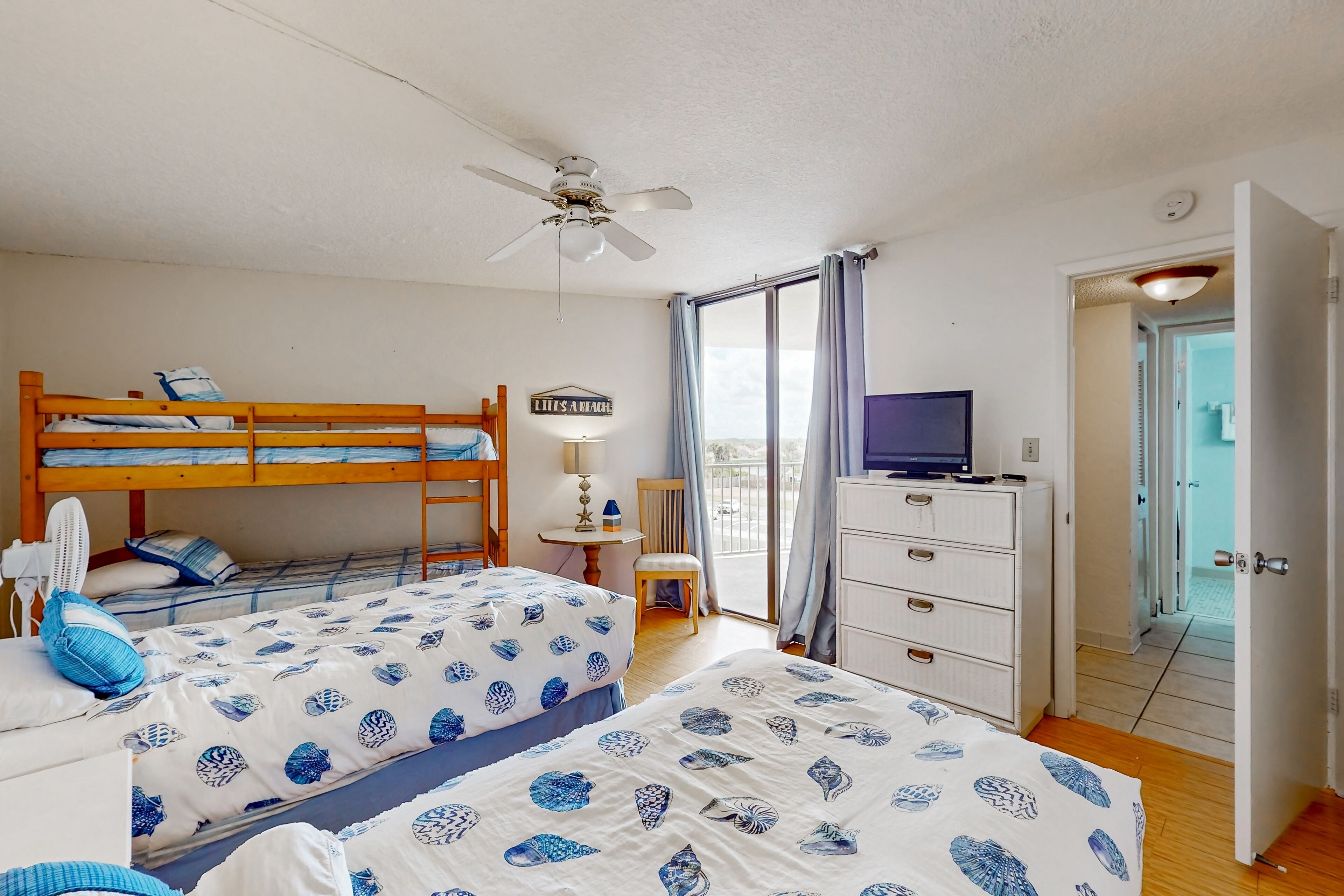 Une chambre avec deux lits, un ventilateur de plafond, une télévision, une commode et une pancarte sur laquelle on peut lire : « LITTS A BEACH ».