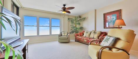 A spacious living room with a large window, a ceiling fan, a sofa, a chair, and a TV.