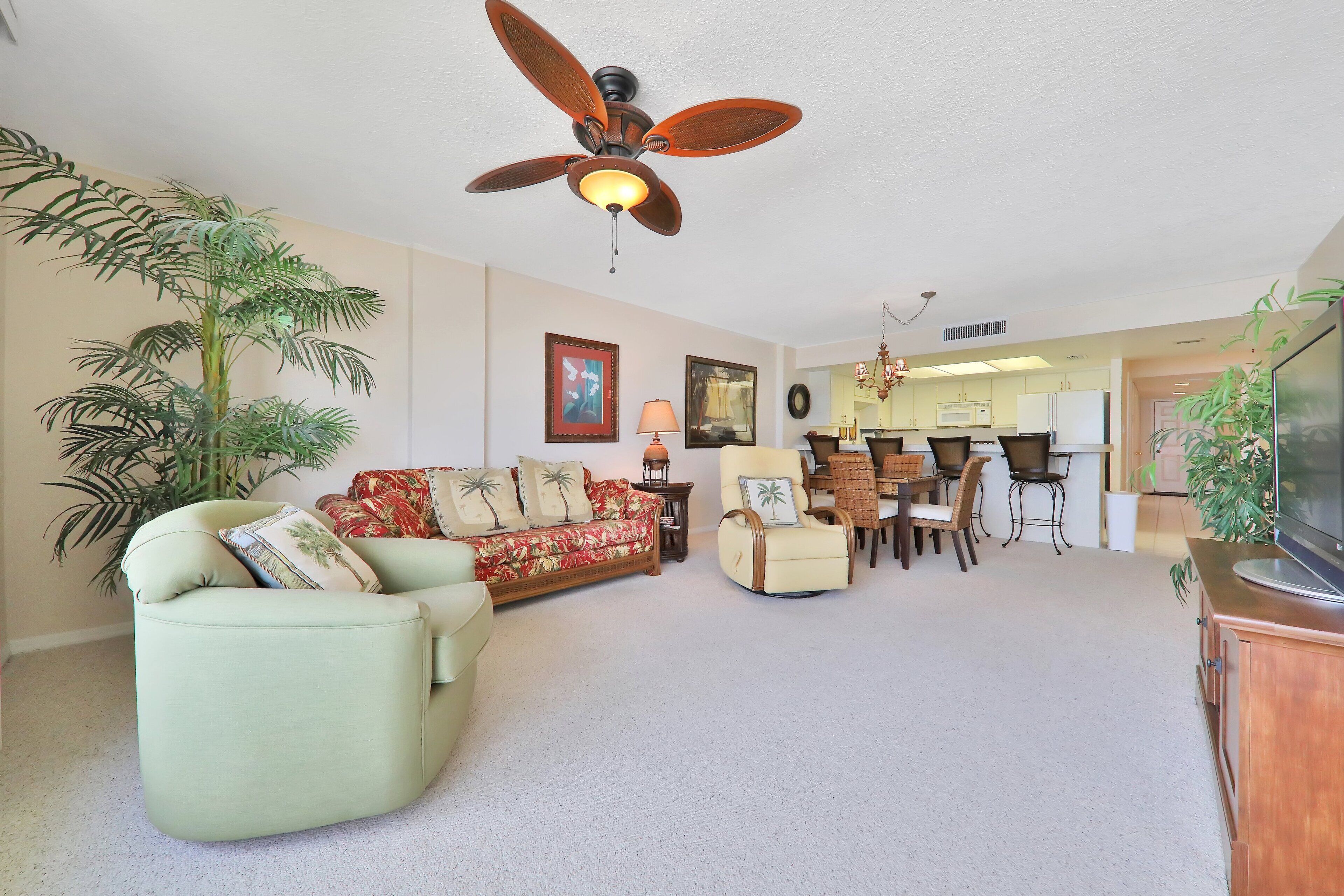 Ein Wohnzimmer mit Sofa, Sessel und Deckenventilator. Ein Essbereich mit Tisch und Stühlen ist ebenfalls vorhanden. Im Hintergrund ist eine Küche zu sehen.