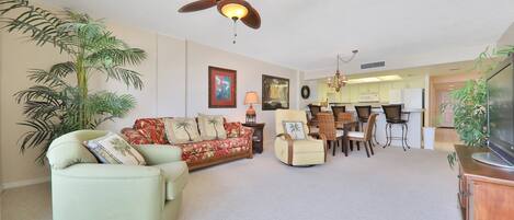 Ein Wohnzimmer mit Sofa, Sessel und Deckenventilator. Ein Essbereich mit Tisch und Stühlen ist ebenfalls vorhanden. Im Hintergrund ist eine Küche zu sehen.