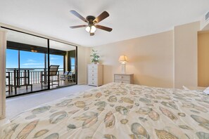 Une chambre avec un grand lit, un ventilateur de plafond, une table de chevet avec une lampe, une commode et une porte-fenêtre coulissante donnant sur un balcon équipé de chaises et d’une table.