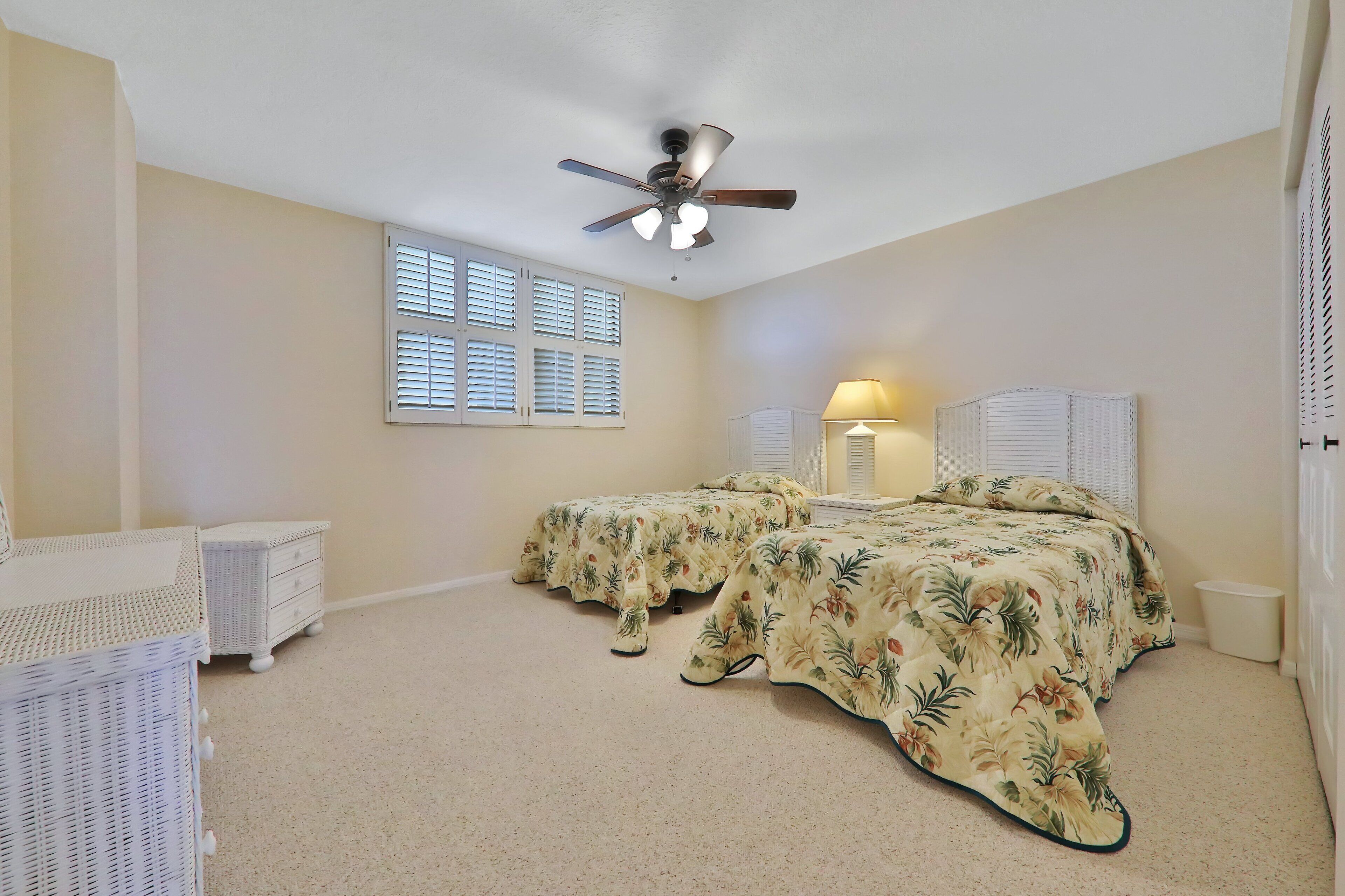 A bedroom with two beds, a ceiling fan, a window with blinds, a bedside lamp, and a wicker chair.