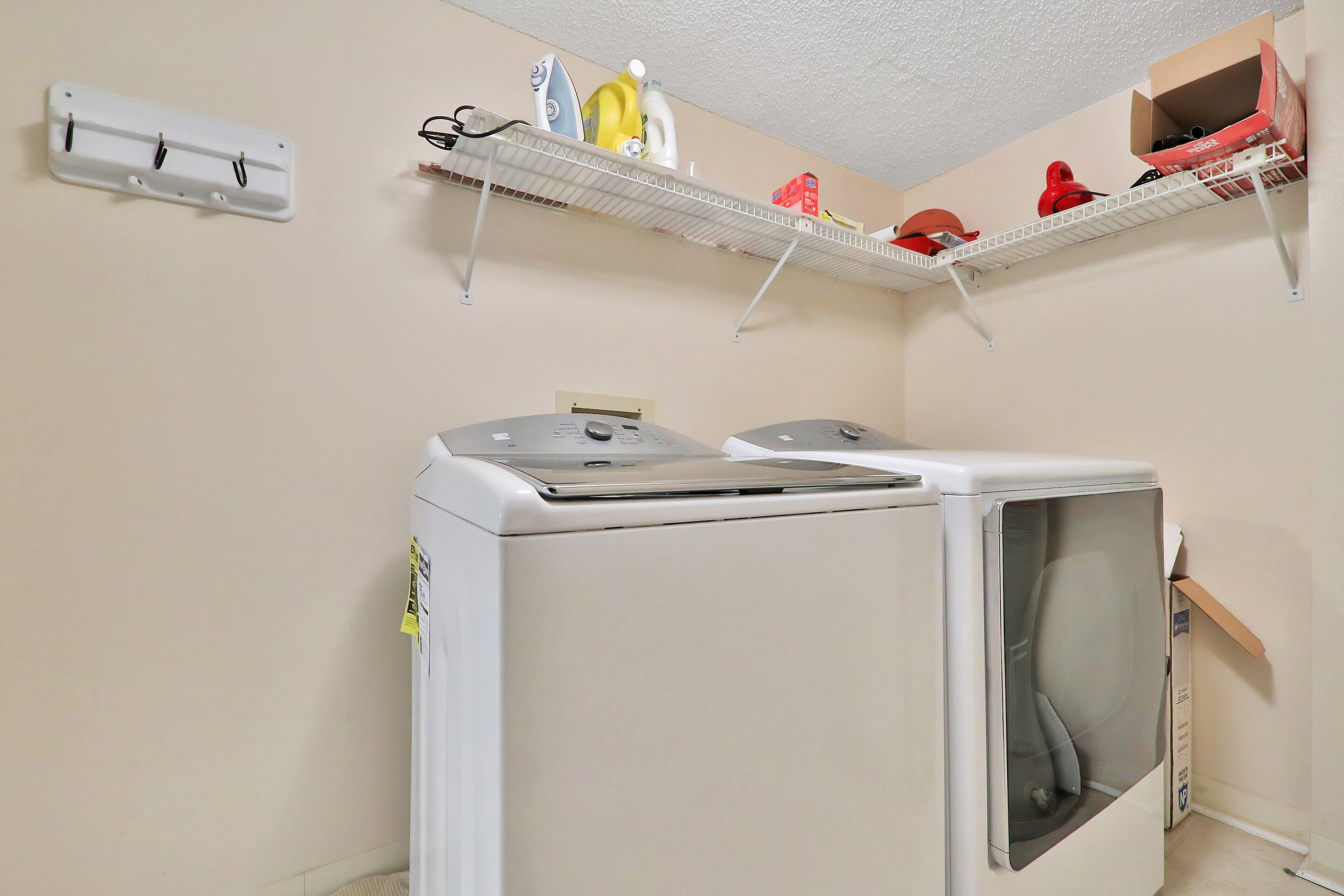 Ett tvättrum med en tvättmaskin med frontmatning, en väggmonterad torktumlare och hyllor ovanför för att förvara olika saker.