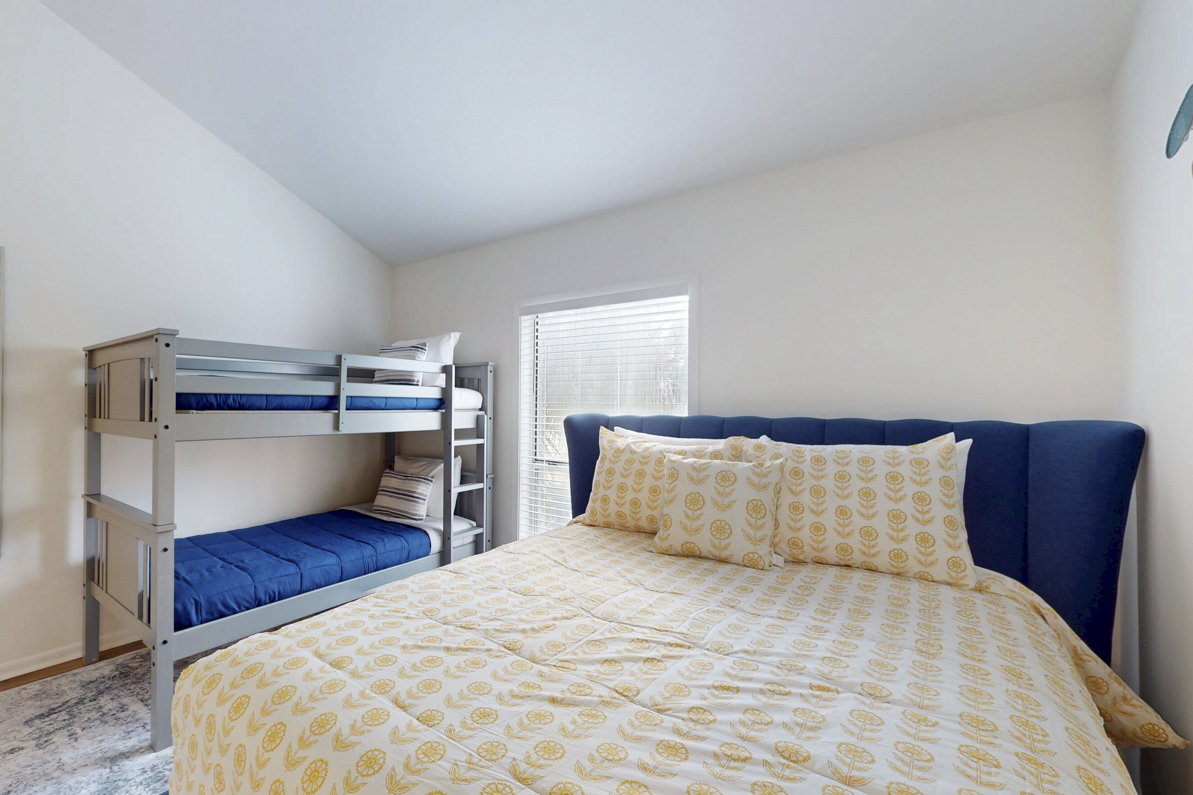 A bedroom with a bunk bed, a single bed with a patterned duvet, and a window with blinds.