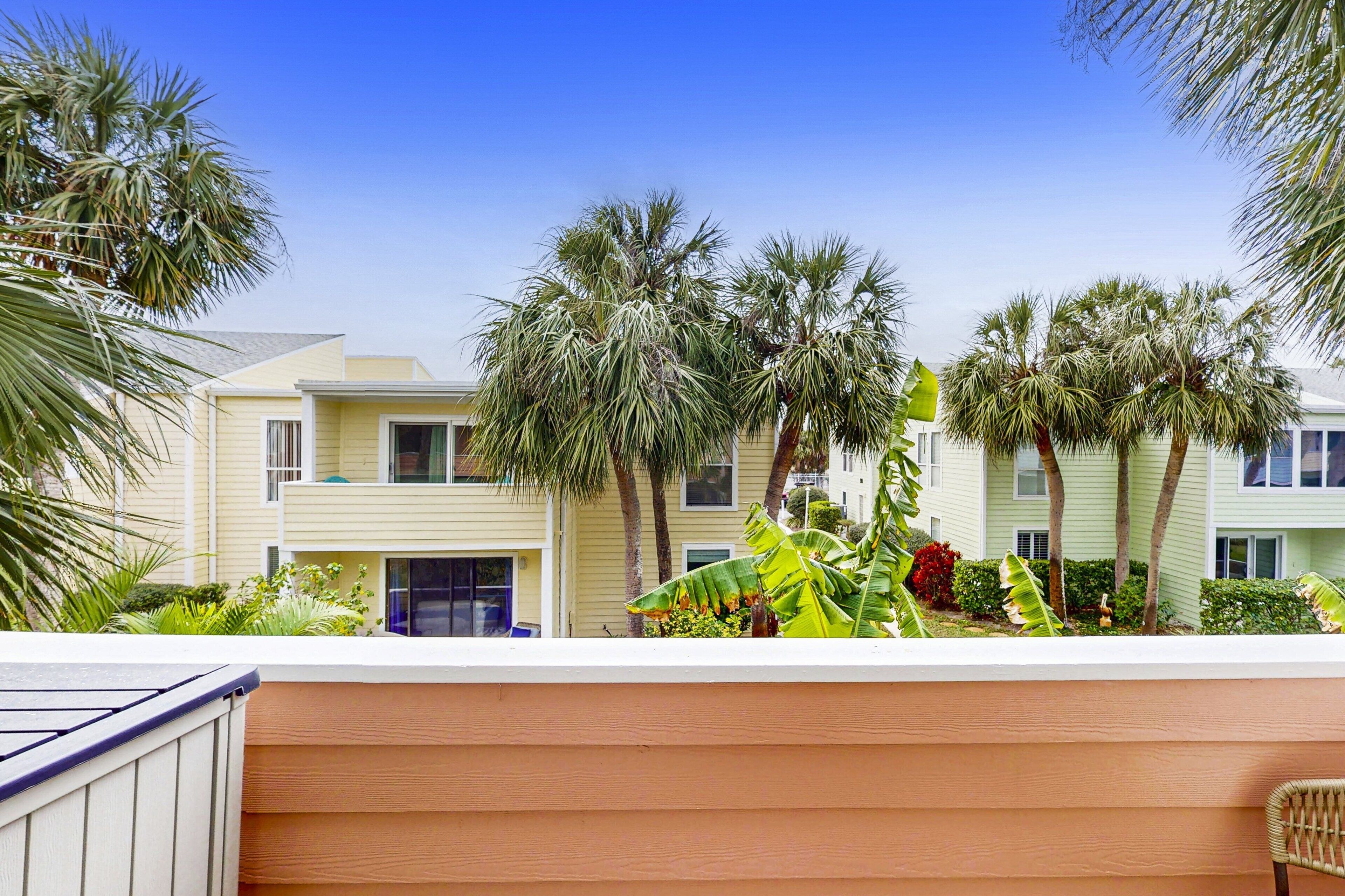 A row of light-colored, two-story buildings with balconies, surrounded by palm trees and tropical plants.