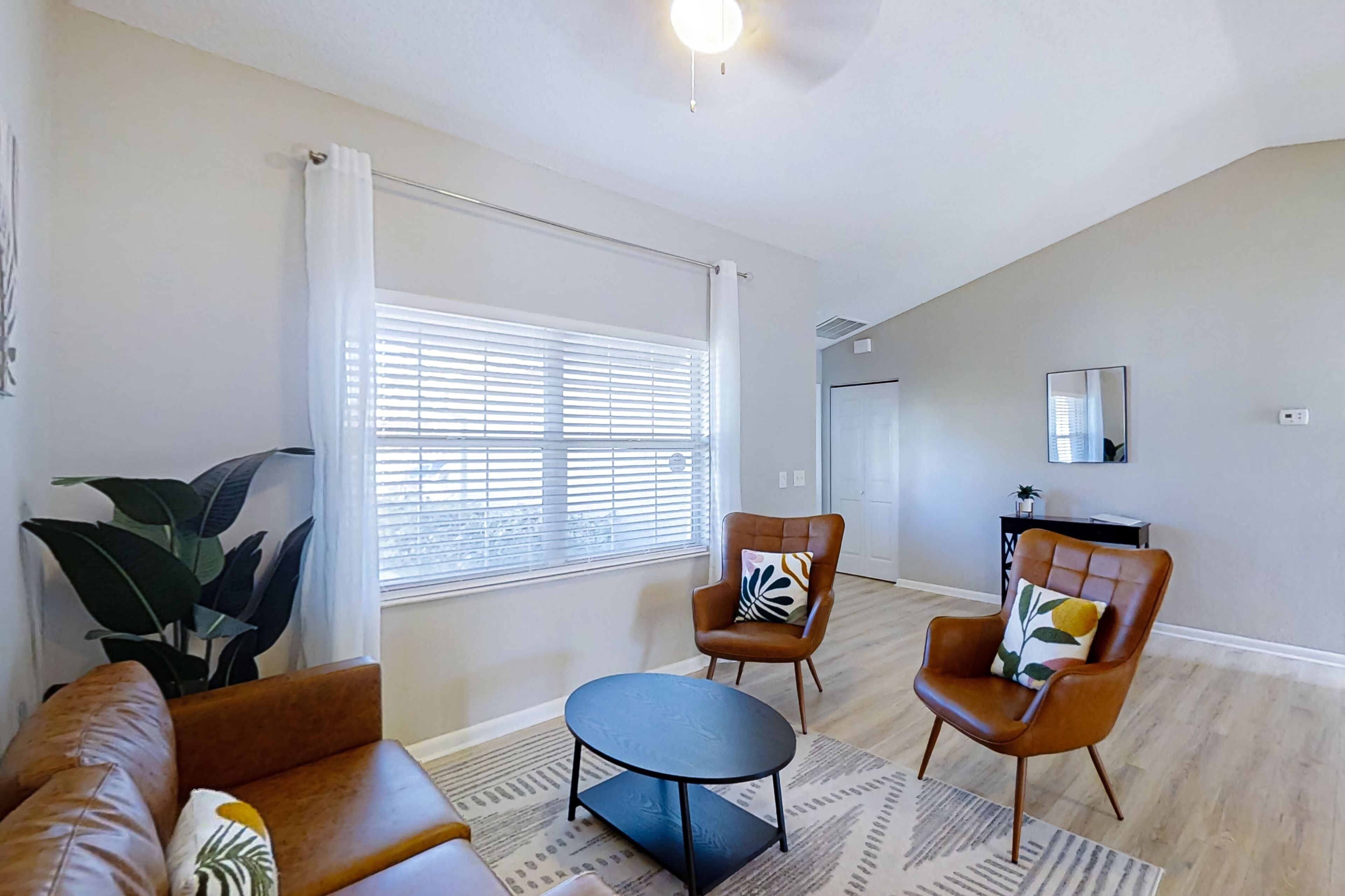 A living room with a brown leather sofa, a round coffee table, and two armchairs with decorative pillows. A large window with blinds allows natural light into the room.