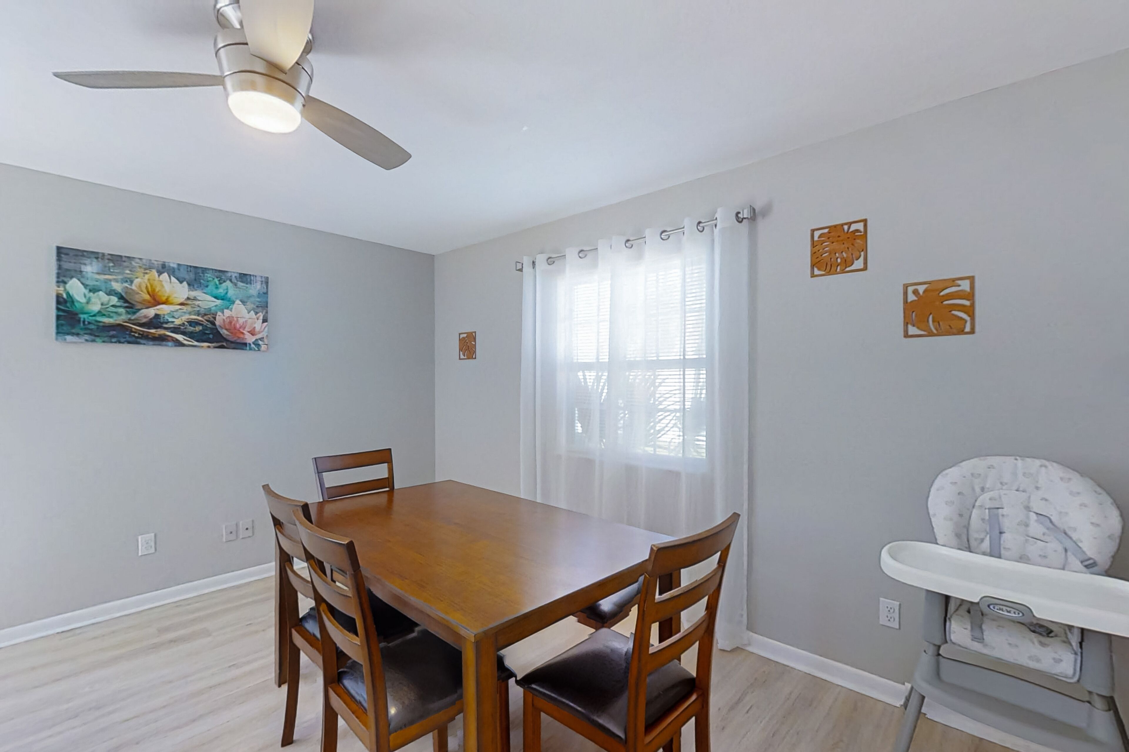 A dining area with a wooden table and chairs, a ceiling fan, a window with sheer curtains, and a baby high chair.