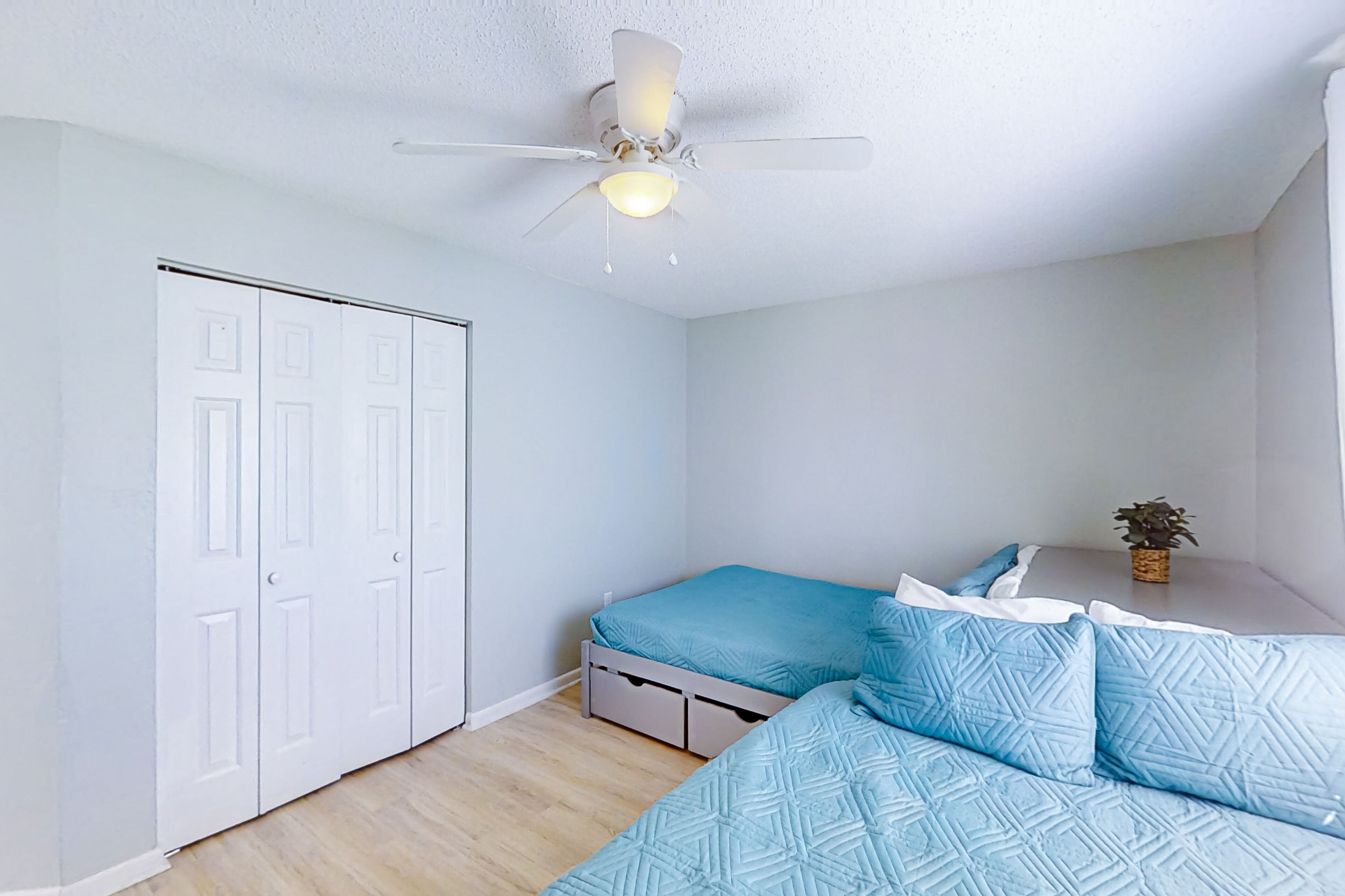 A bedroom with a bed, a ceiling fan, a wardrobe, and a small table with a plant.