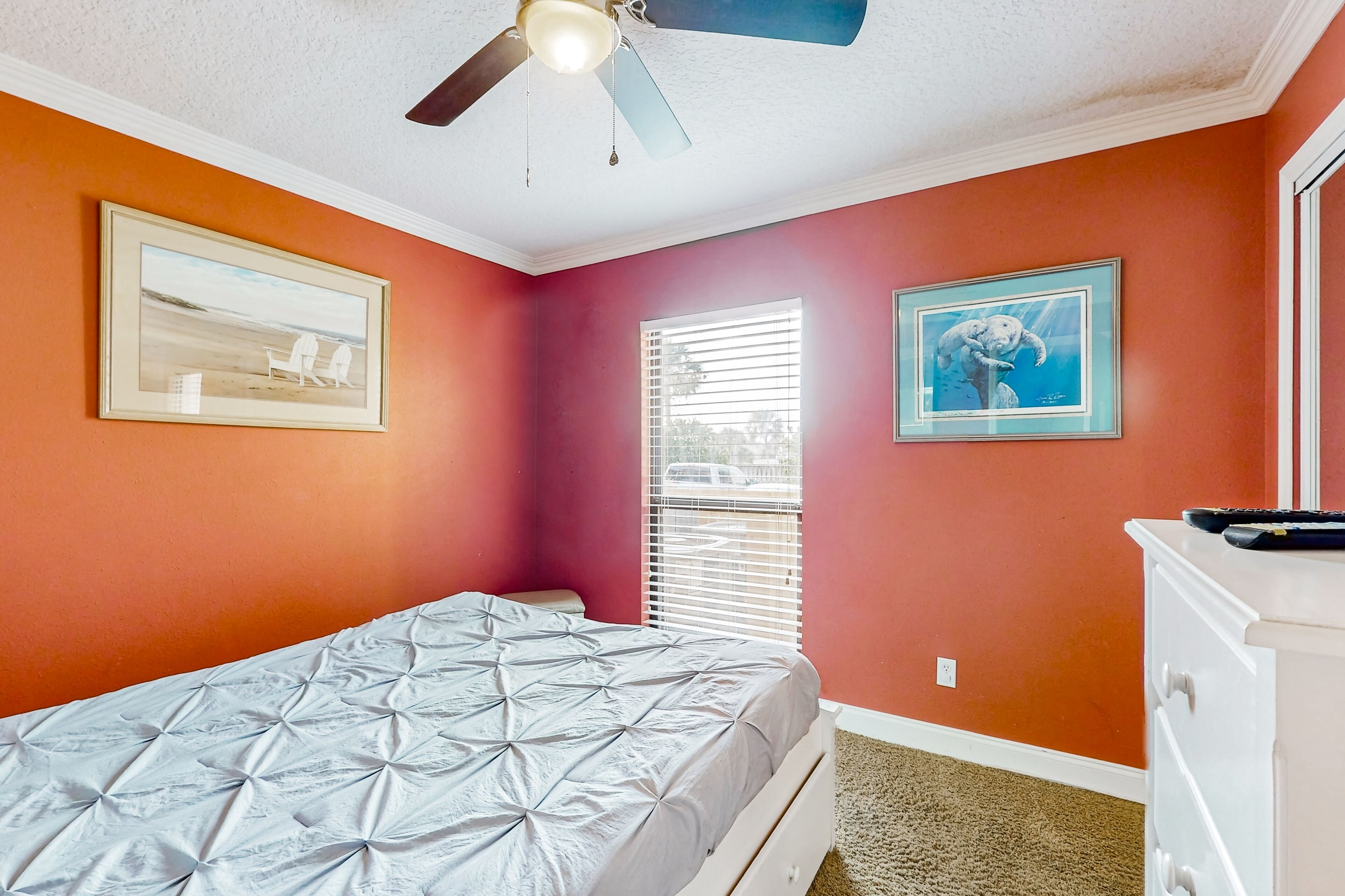 A bedroom with an orange wall, a bed with a grey bedspread, a ceiling fan, and two framed pictures.