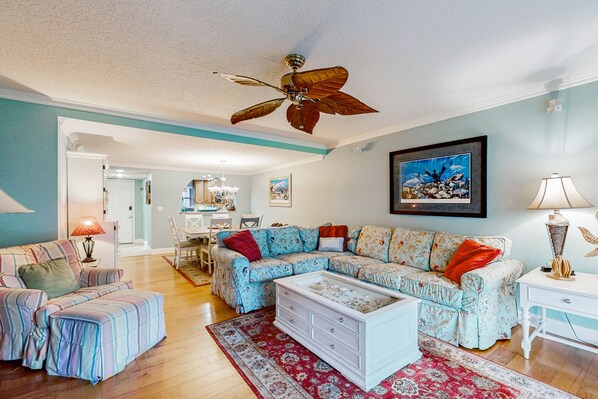 A living room with a sectional sofa, ottoman, and a coffee table. There is a ceiling fan, a chandelier, and a framed picture on the wall. - Patio Condo in Oceanfront Community w/ Pool Walk to Beach (St. Augustine)