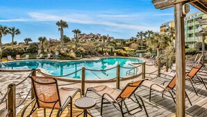Ein Poolbereich mit Liegestühlen und einer Holzterrasse, umgeben von Palmen und Gebäuden im Hintergrund.