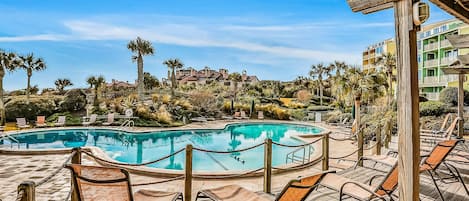 A pool area with lounge chairs and a wooden deck, surrounded by palm trees and buildings in the background.