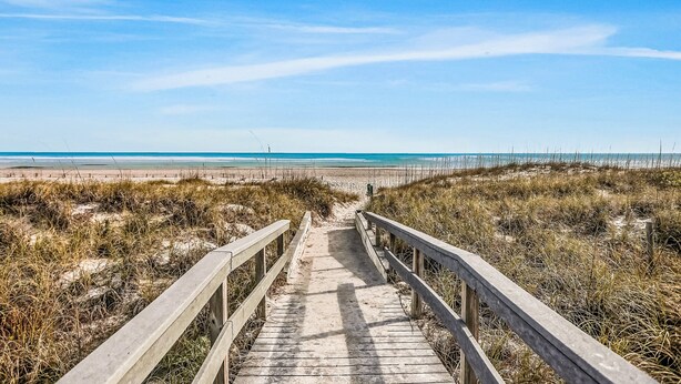 Ein hölzerner Steg führt zu einem Strand unter blauem Himmel.