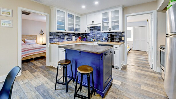 A compact kitchen with a central island, bar stools, and a bed in the adjacent room. - Sleek Golf-front Condo With a Washer/dryer, Deck, & Central AC - dog Friendly (St. Simons Island)