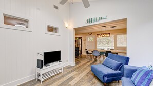 A living room with a TV, blue sofas, a dining area with a table and chairs, and a kitchen in the background. - Sleek Golf-front Condo With a Washer/dryer, Deck, & Central AC - dog Friendly (St. Simons Island)