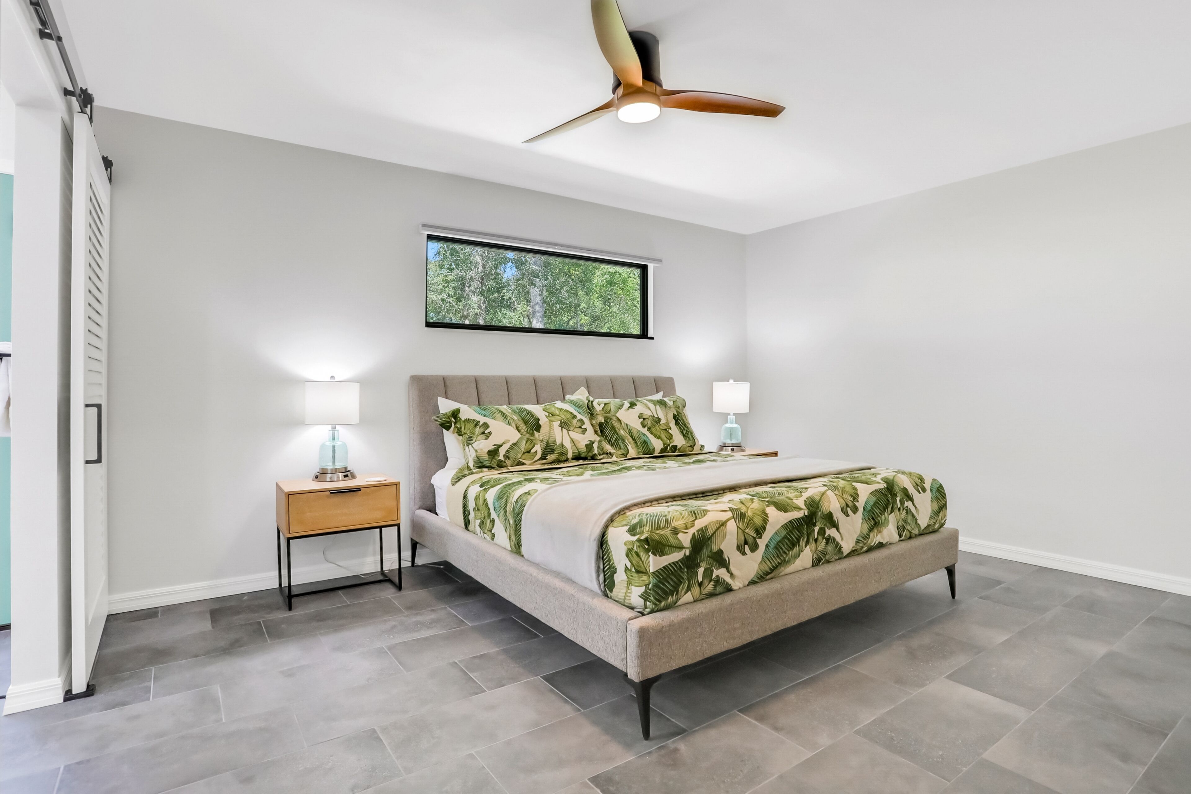 Quarto com cama, mesas de cabeceira, ventilador de teto e janela com vista para árvores.