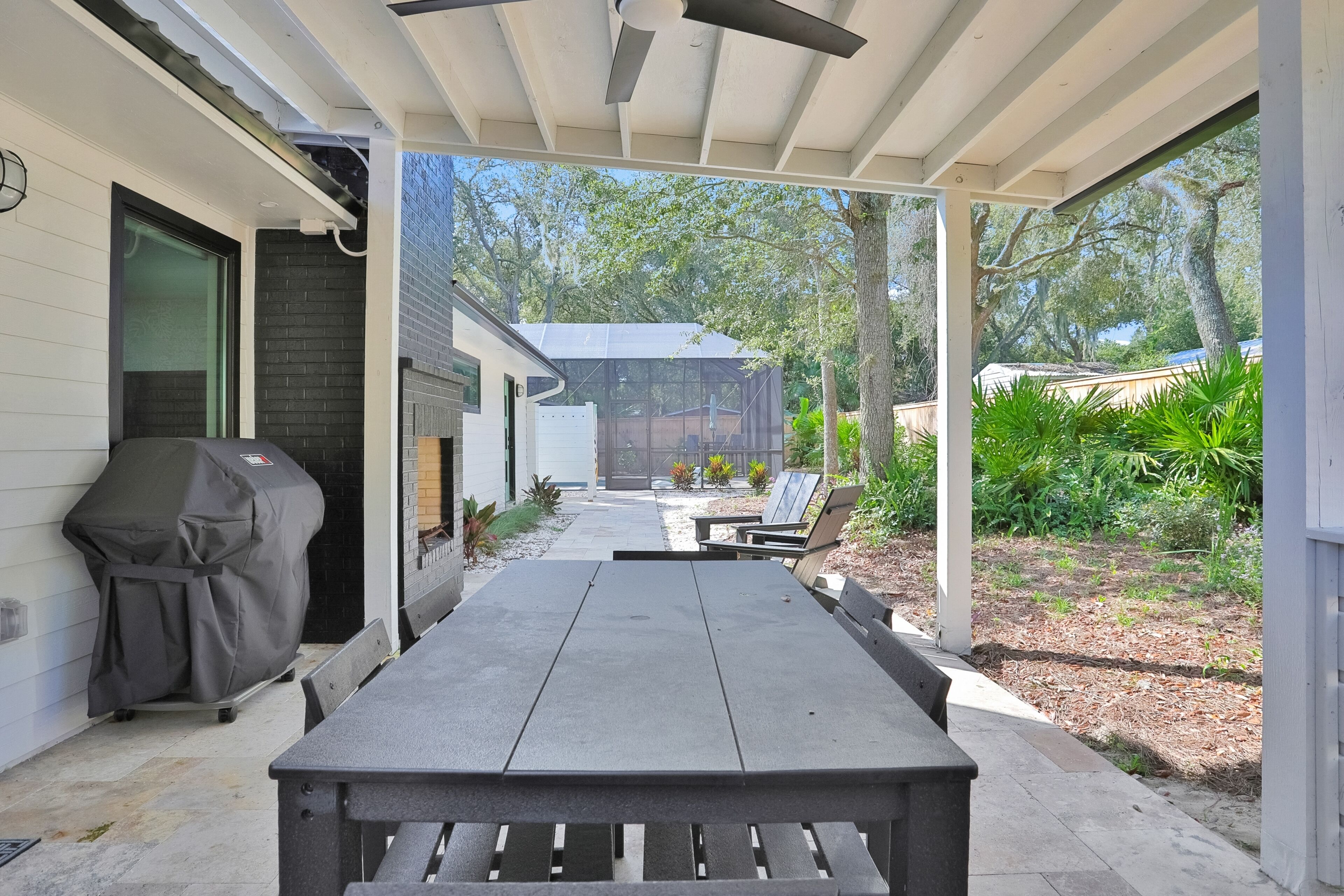 An outdoor patio with a covered table and chairs, a barbecue grill, and a view of a house and garden.