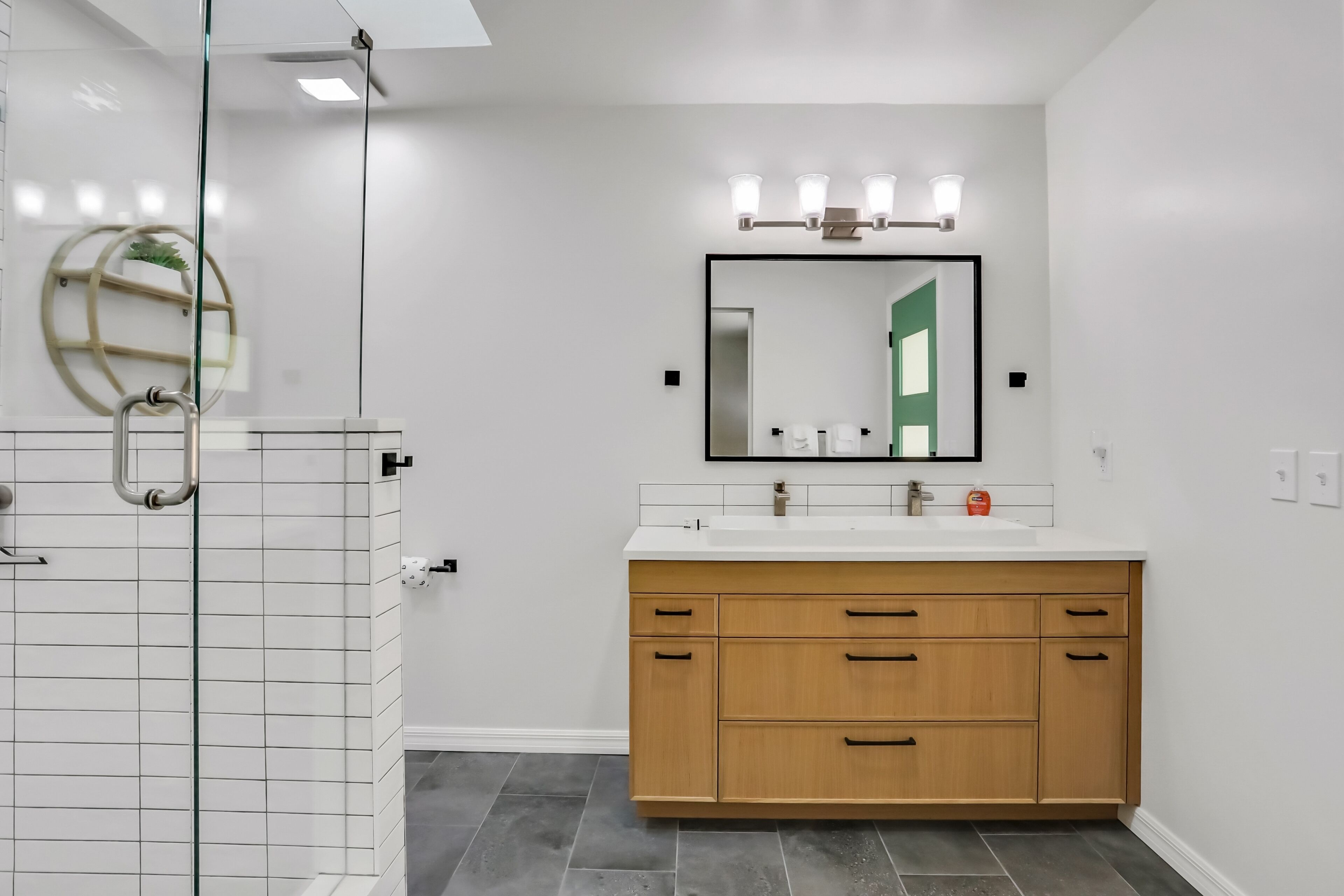 A modern bathroom with a glass shower enclosure, a wooden vanity with a large mirror, and a white tiled floor.