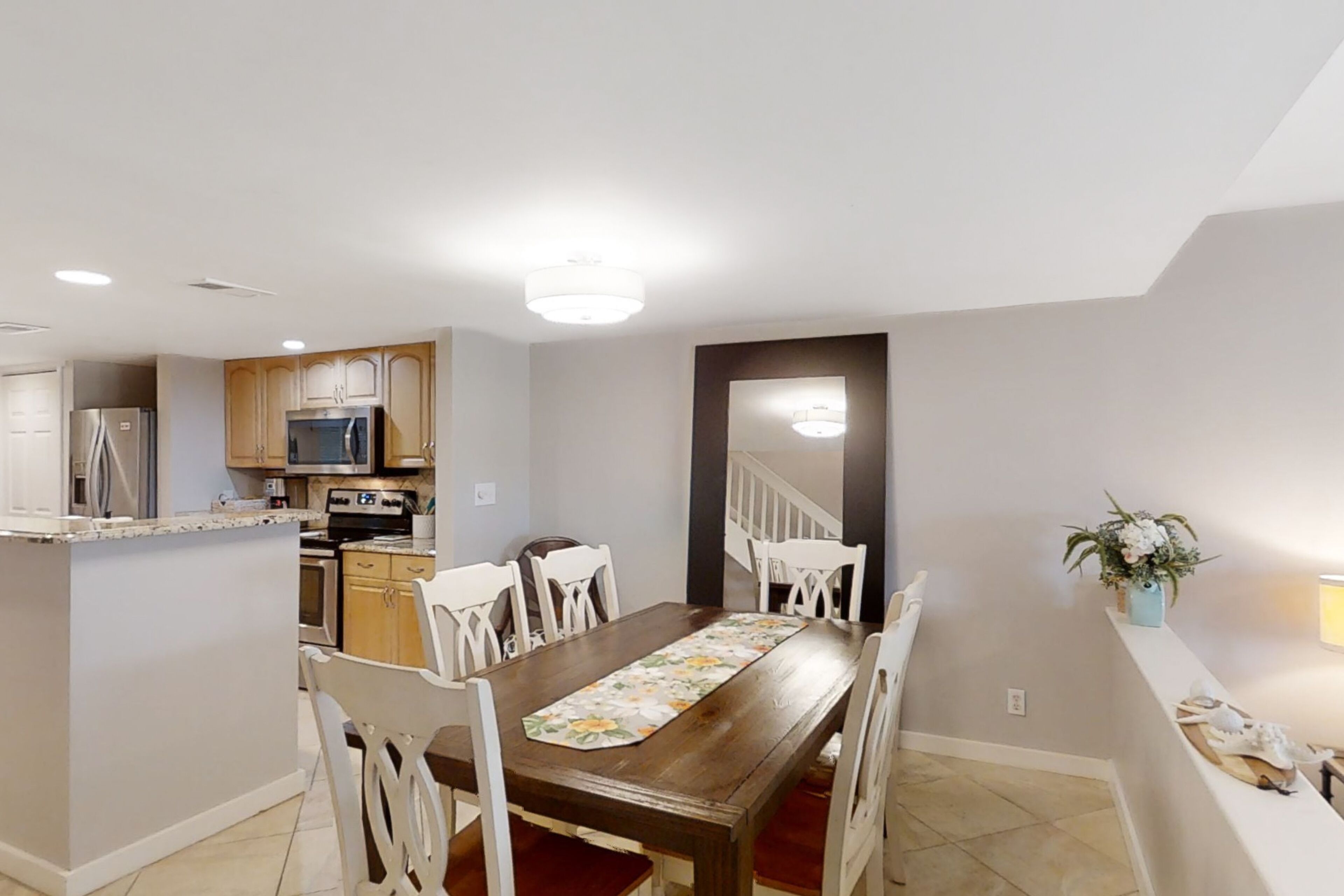 Un comedor con mesa de madera y sillas blancas, una cocina con electrodomésticos modernos y una escalera que conduce a otro nivel.