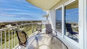 A balcony with a glass table and four chairs, overlooking a coastal landscape with a bridge and greenery. - Oceanfront, 2nd-floor Condo With Balcony, Hdtvs, Wifi, AC & Shared Pool (Fernandina Beach)