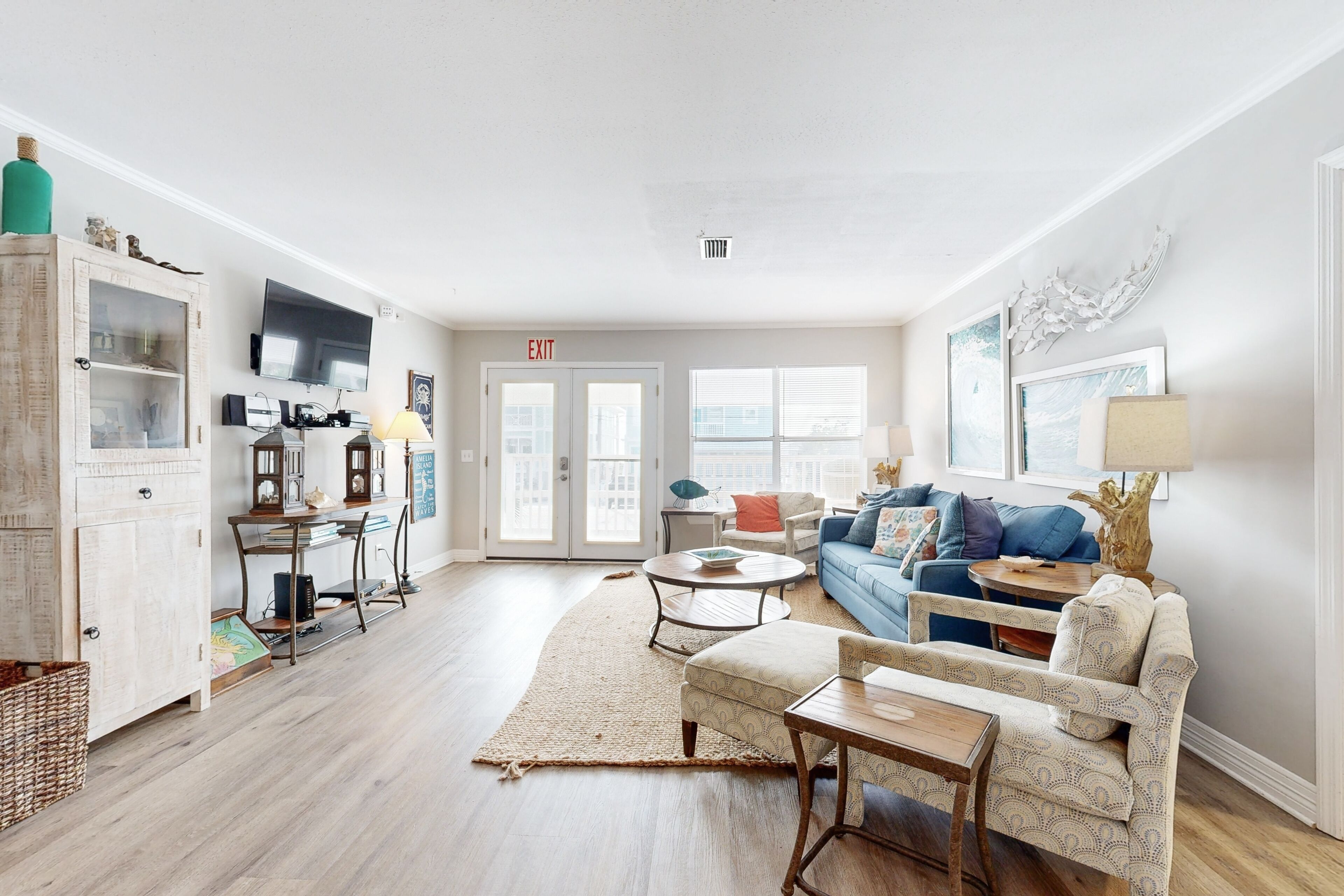 A spacious living room with a blue sofa, a wooden coffee table, and a white armchair. There is a bookshelf with decorative items and a flat-screen TV mounted on the wall.