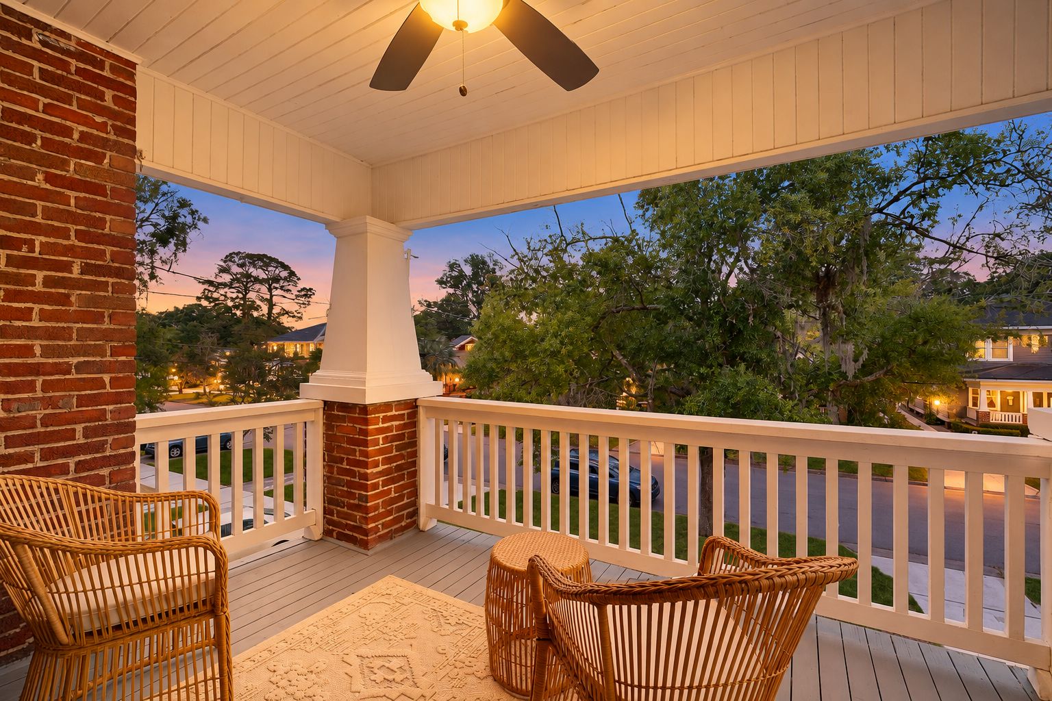 A covered porch with wicker furniture and a brick chimney, overlooking a residential street at dusk.