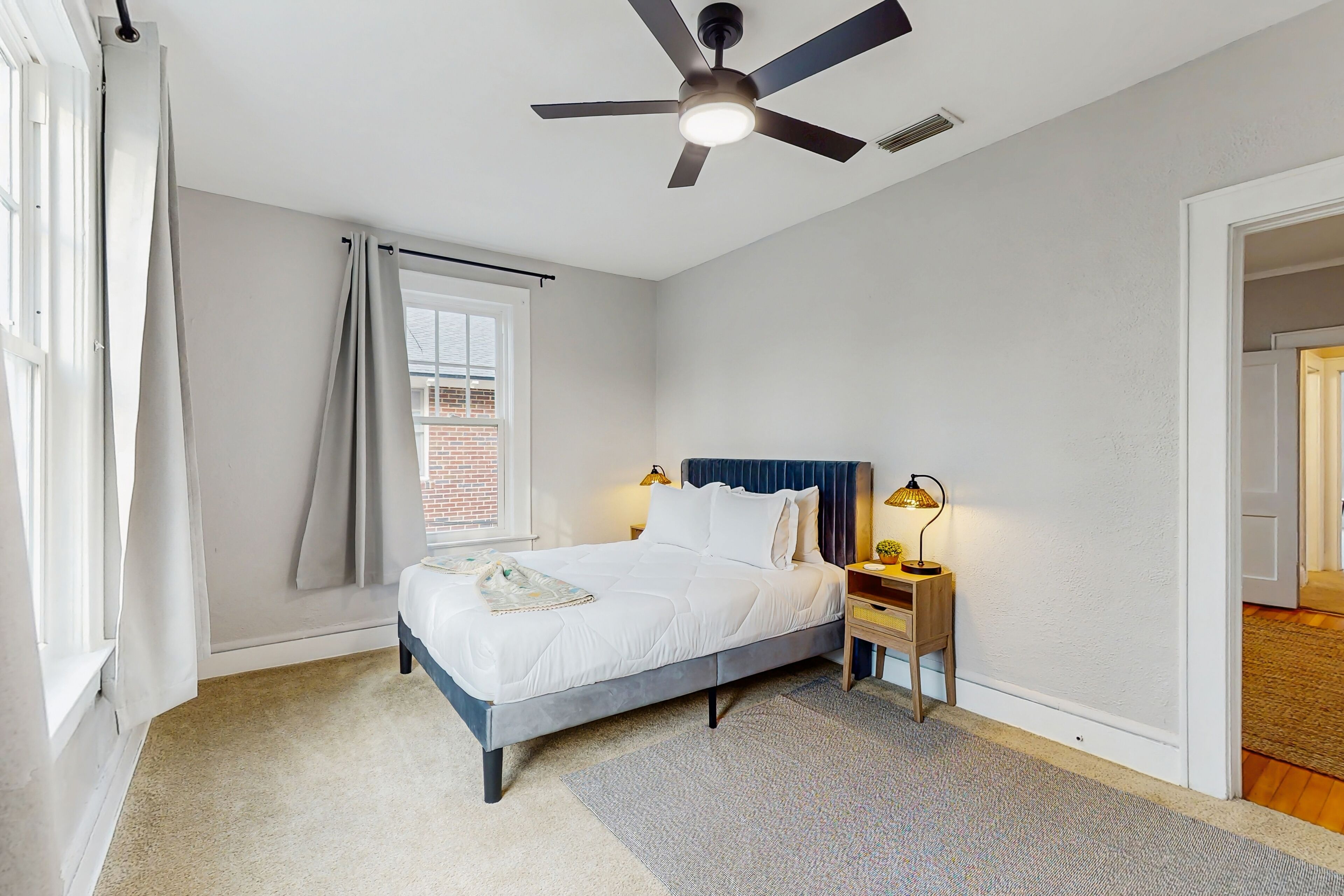 A bedroom with a bed, bedside lamps, a ceiling fan, a window with curtains, and a view of a brick building.