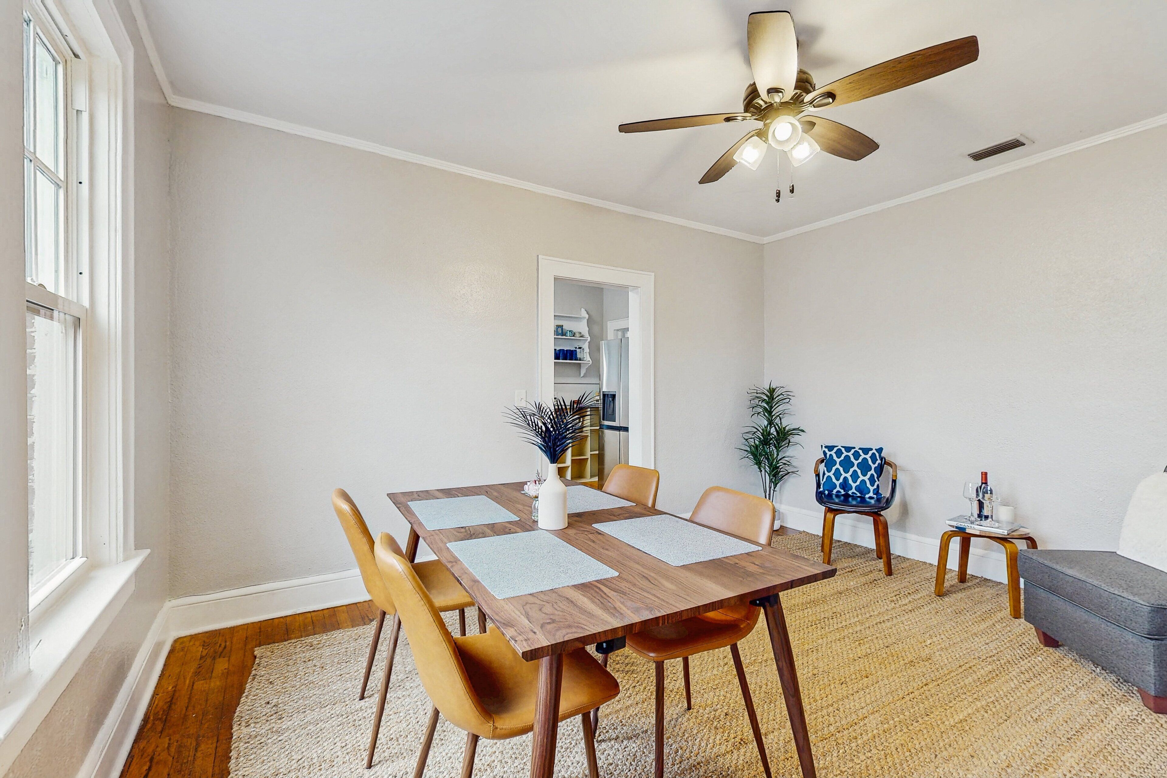 A dining area with a wooden table, four chairs, a ceiling fan, and a view of a kitchen through an open doorway.
