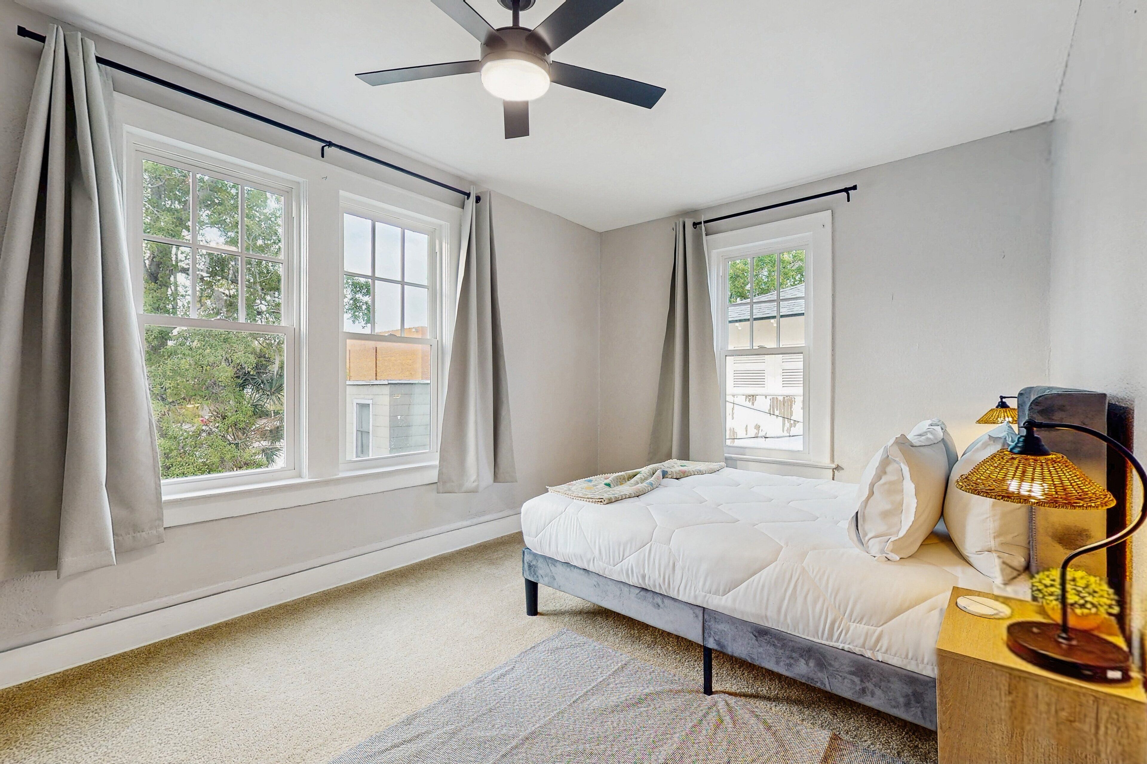 A bedroom with a bed, a ceiling fan, a nightstand with a lamp, and large windows with curtains.