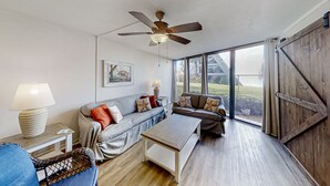 A spacious living room with a ceiling fan, a sofa, a coffee table, and a view through large windows. - Renovated, Ground-floor Condo at Oceanfront Complex With Pool - Steps to Beach (St. Augustine)