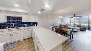 A modern kitchen with white cabinets, a blue-tiled backsplash, and a large island. There is a dining area with a wooden table and chairs, and a sofa in the living room. - Renovated, Ground-floor Condo at Oceanfront Complex With Pool - Steps to Beach (St. Augustine)
