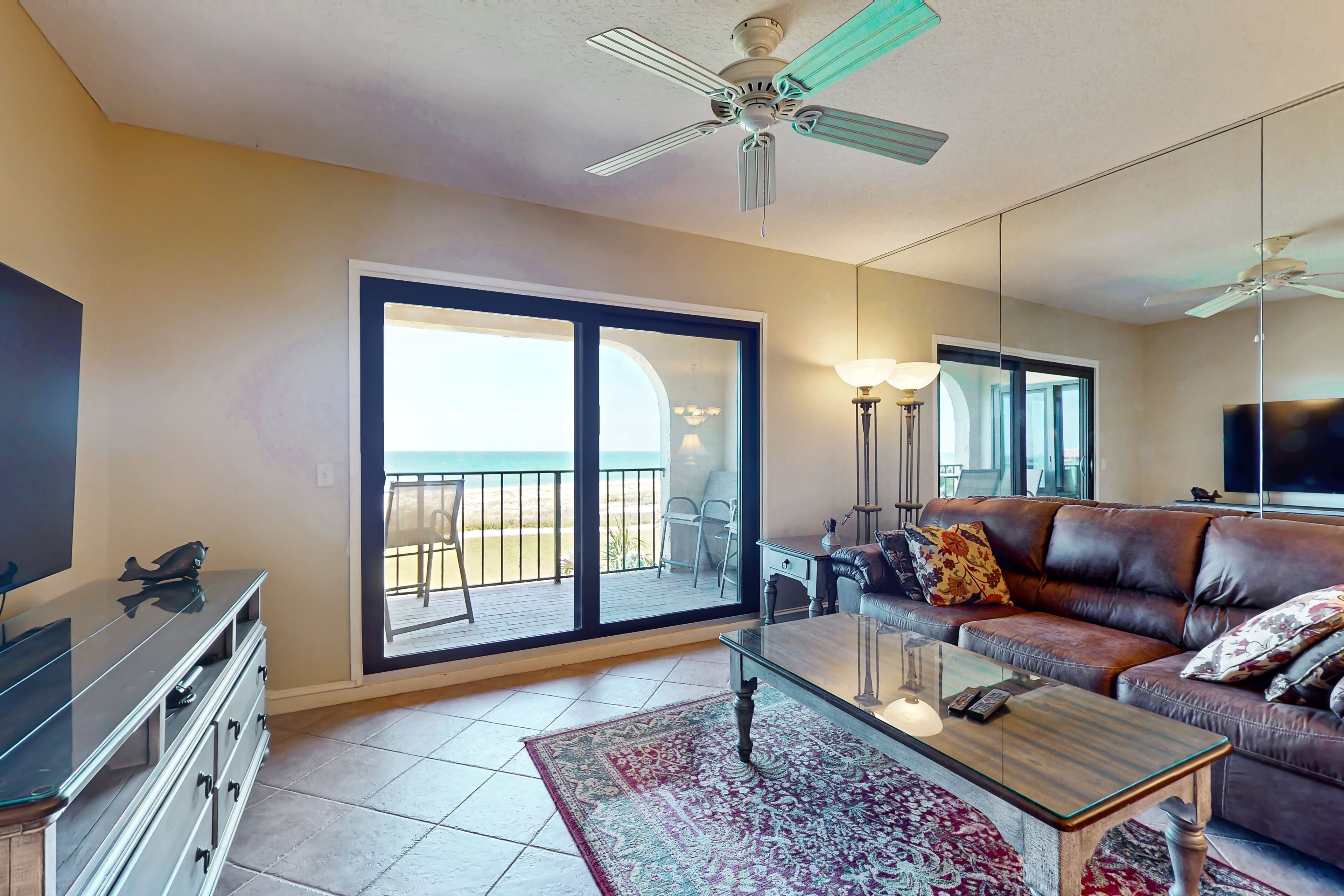 A living room with a glass coffee table, a brown leather sofa, a TV on a stand, and a balcony with a view.