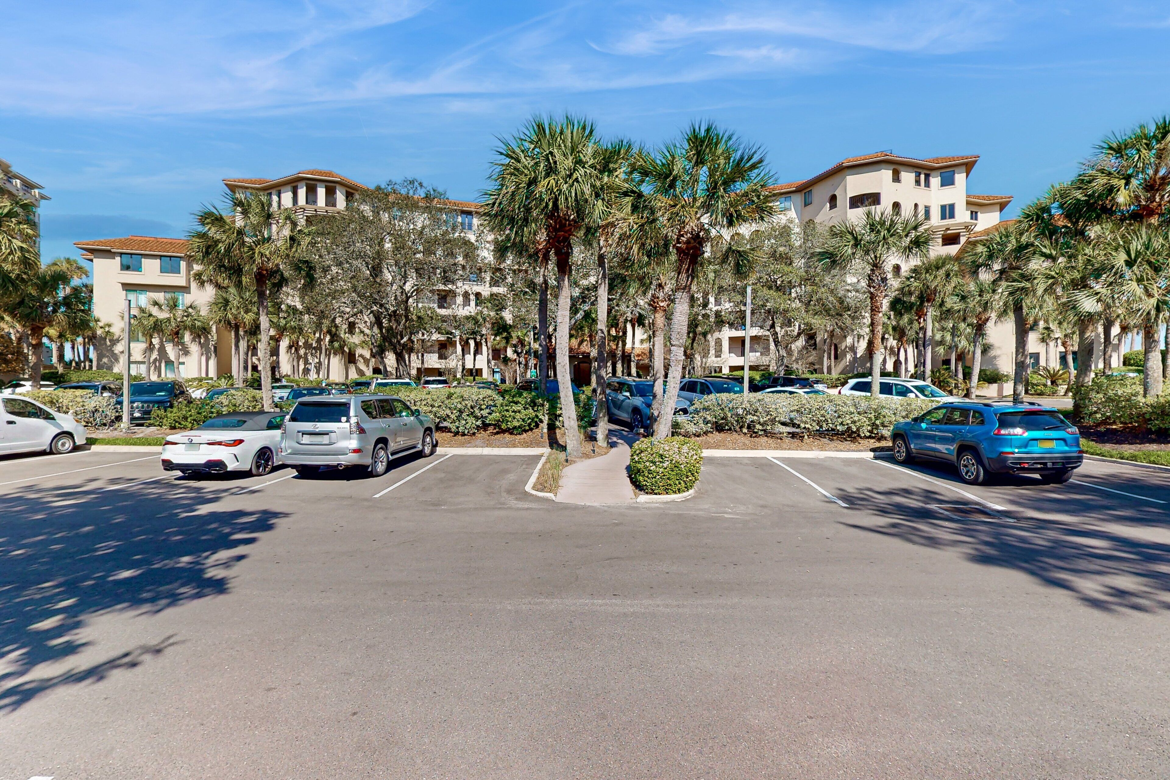 A parking lot with several cars, palm trees, and a building with a tiled roof.