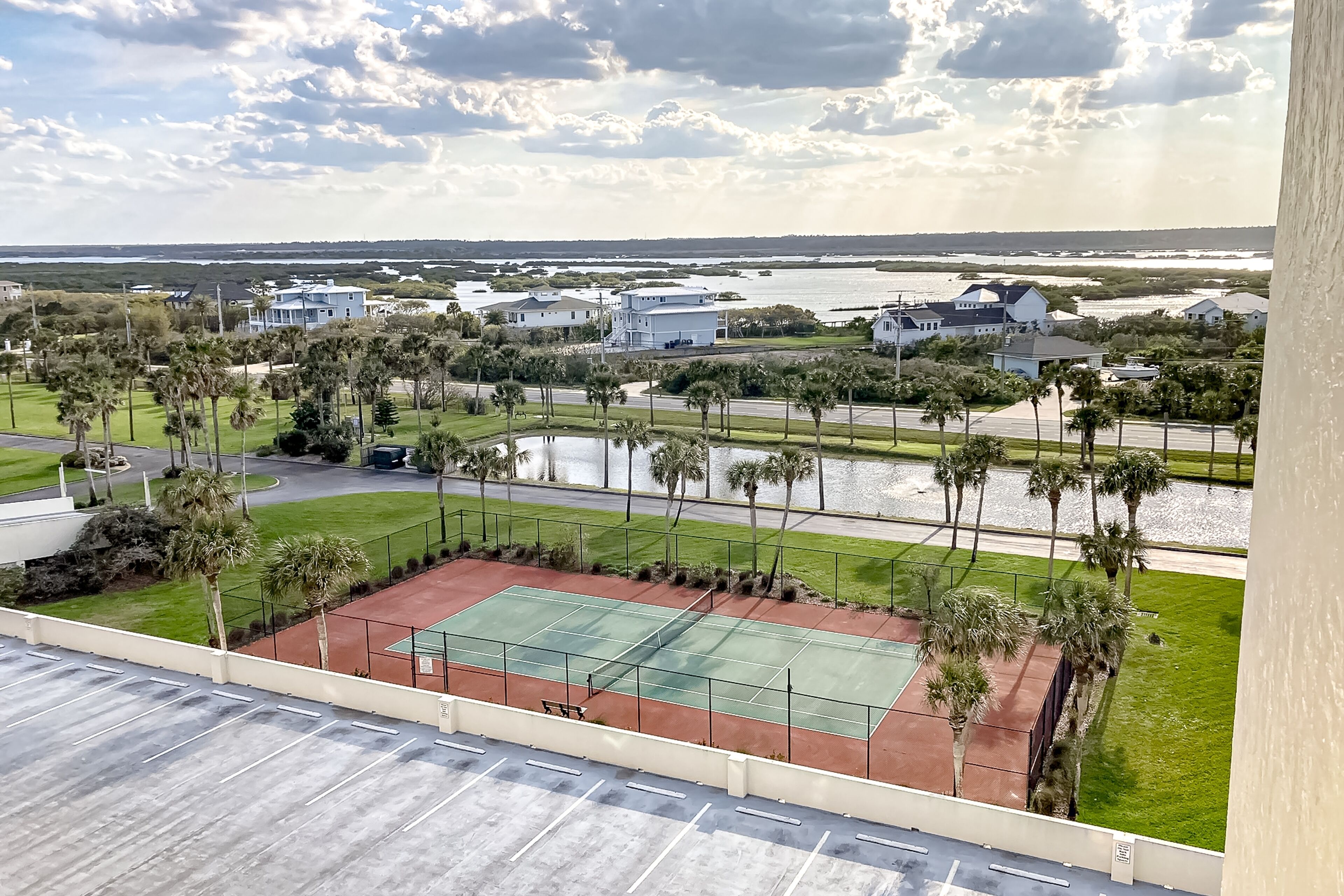 Un court de tennis entouré de palmiers et de maisons, avec vue sur une étendue d’eau et un ciel nuageux.