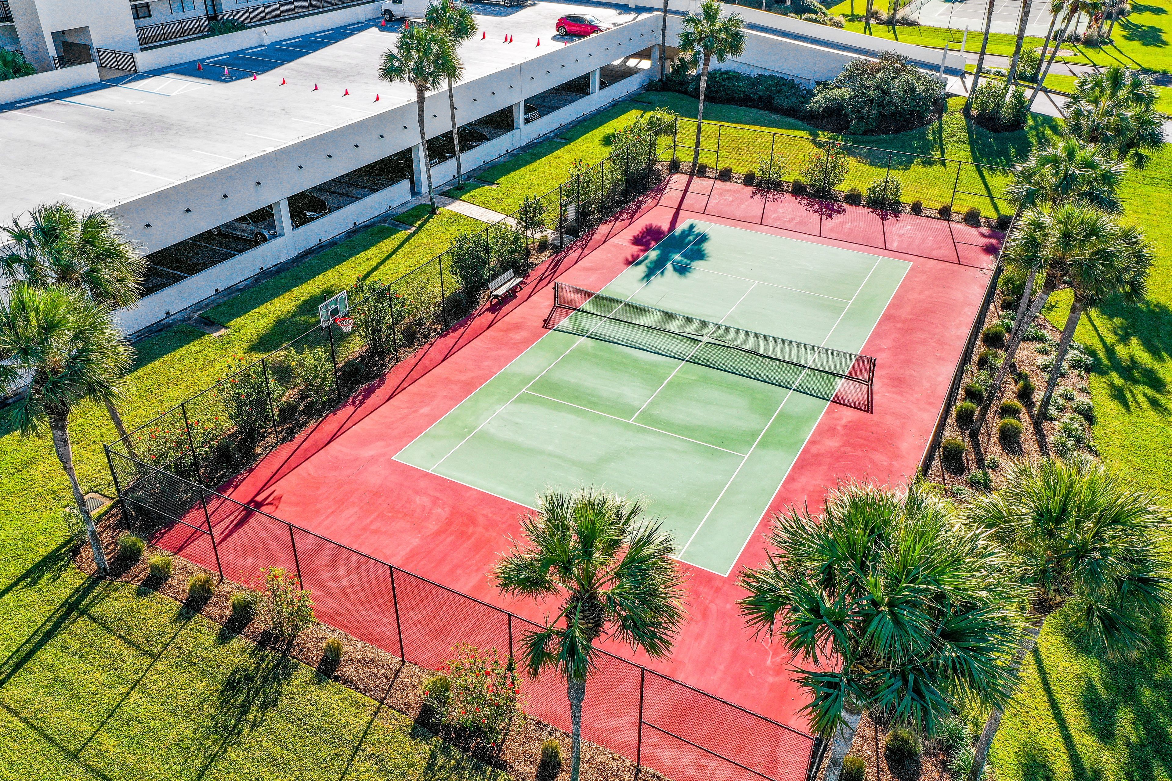 An aerial view of a tennis court surrounded by palm trees and a parking garage.